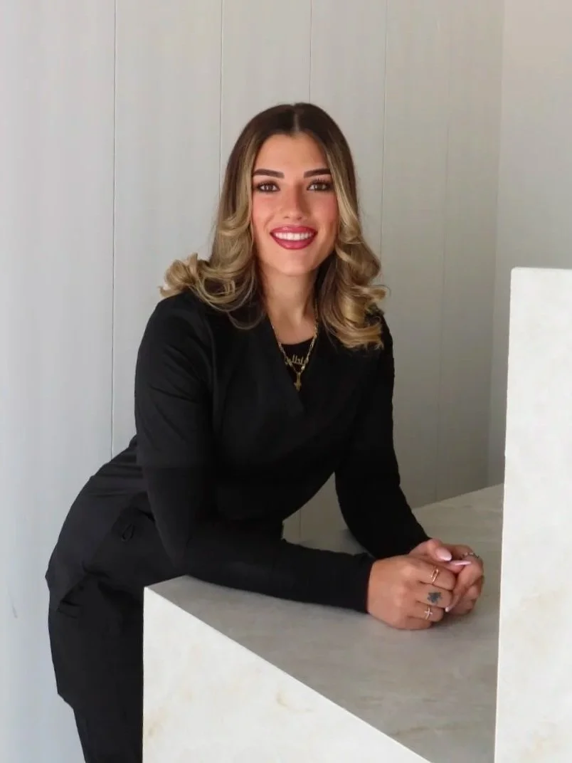 A young woman with shoulder-length wavy hair, smiling, wearing a black long-sleeve top and jewelry, leaning on a white counter in a modern, minimalist room.