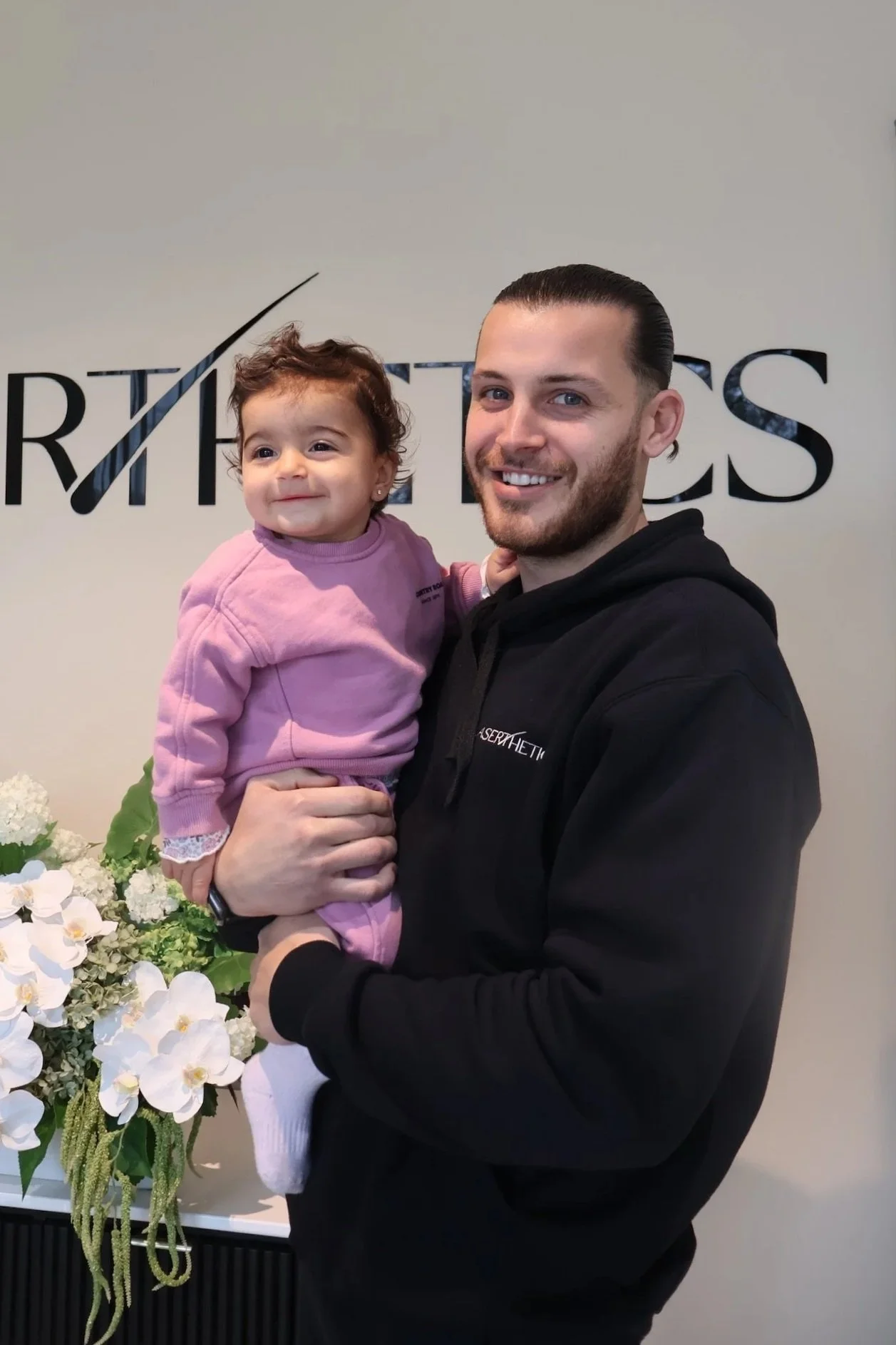 A man with short dark hair and a beard holding a young girl with curly brown hair. They are smiling and standing in front of a wall with the word 'RHTH' and a tilted check mark. There are white flowers placed on a surface behind them.