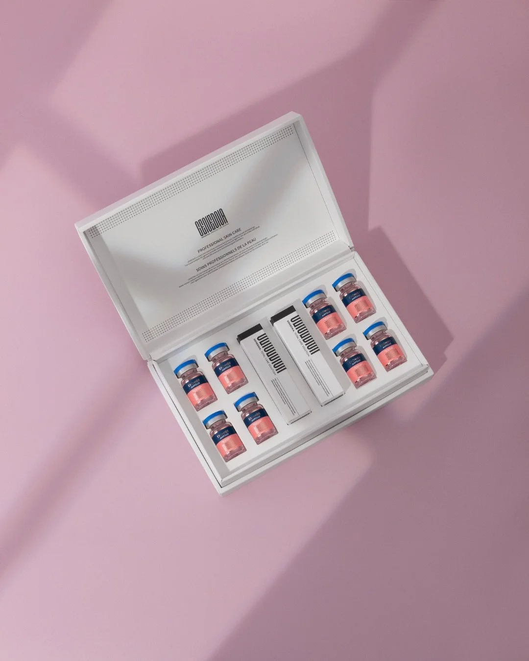 A white box containing small bottles and boxes of skincare products on a pink surface with shadows.