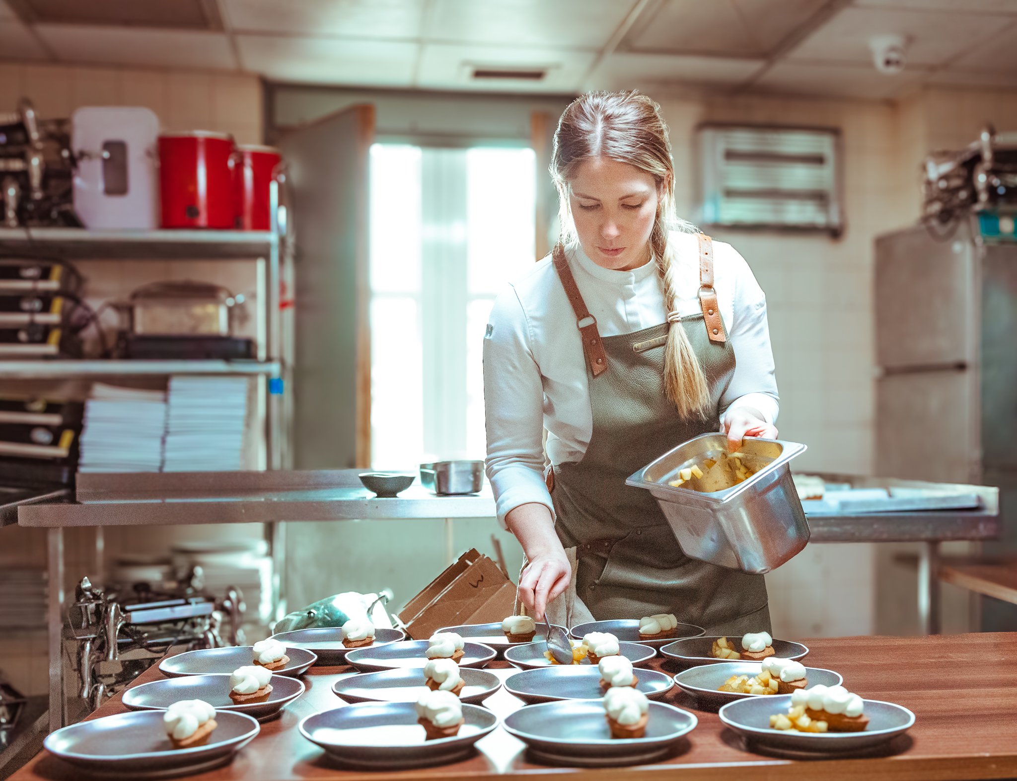 A woman in a kitchen with a brown apron preparing plates with food, such as small desserts and fruit.