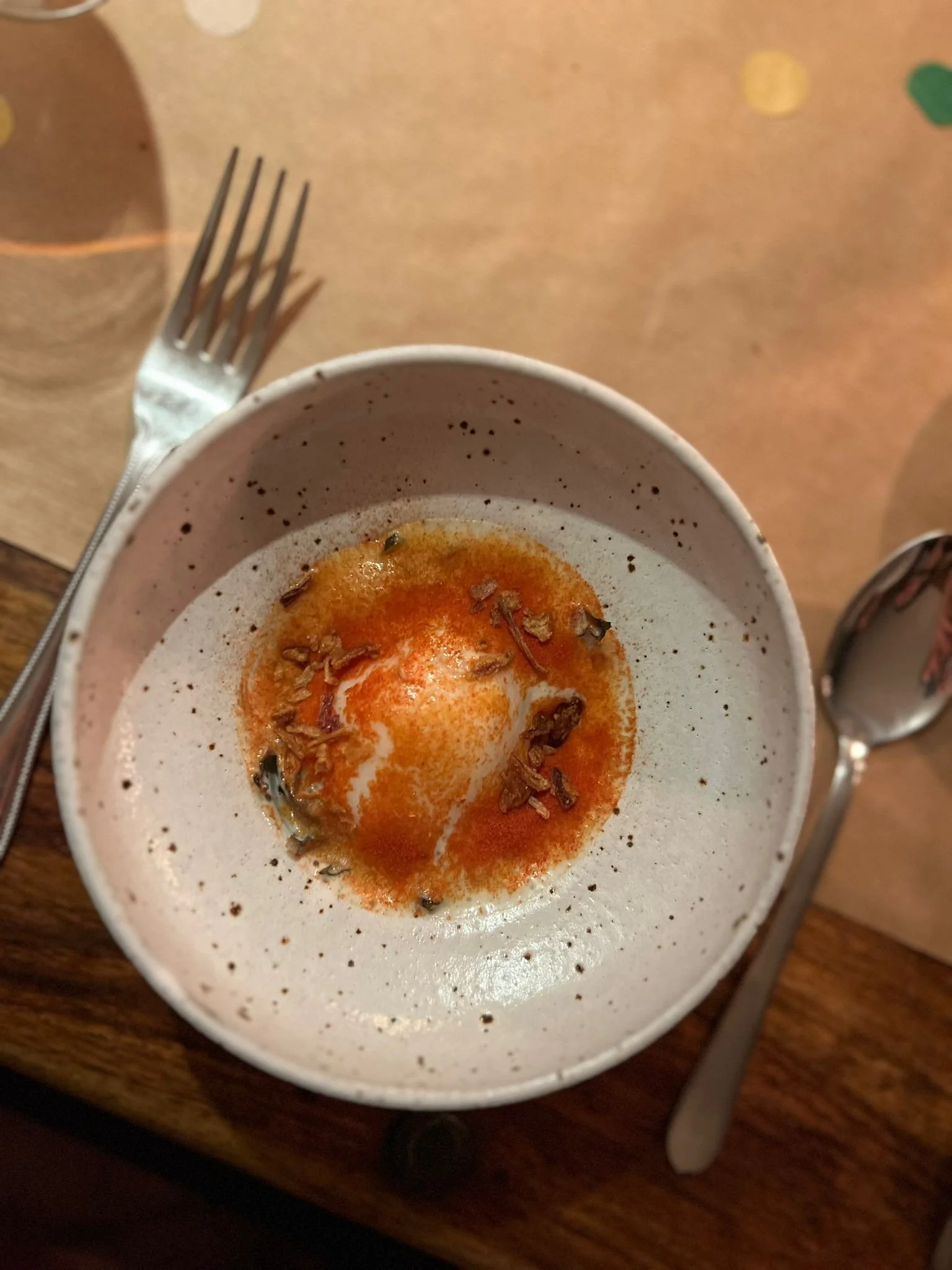 Bowl with a soft-boiled egg topped with chili oil and chopped herbs, placed on a wooden table with a fork and spoon nearby.