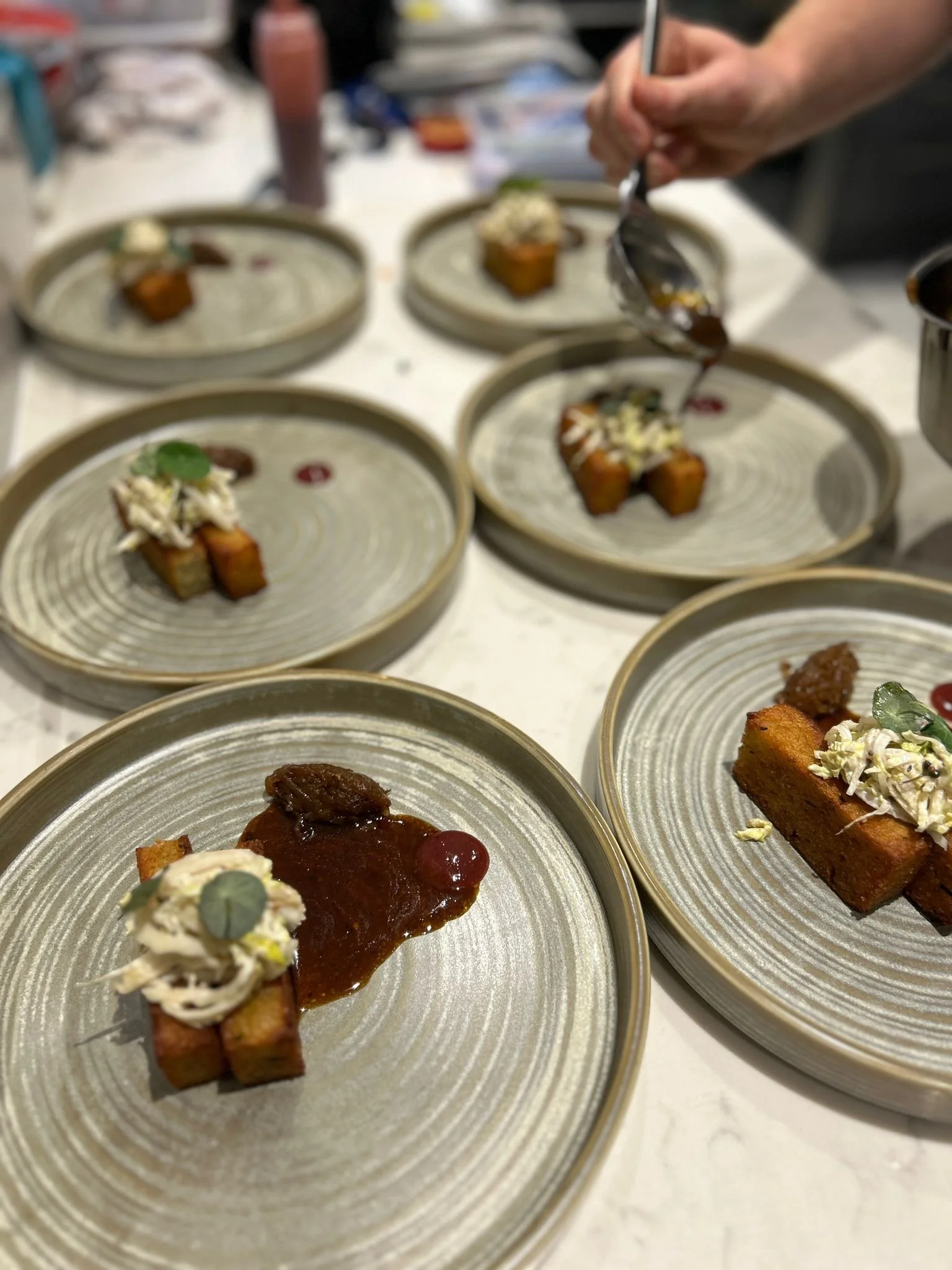 Multiple plated dishes featuring pieces of fried tofu topped with sauce and garnished with herbs, served alongside dollops of creamy sauce and red sauce on ceramic plates.