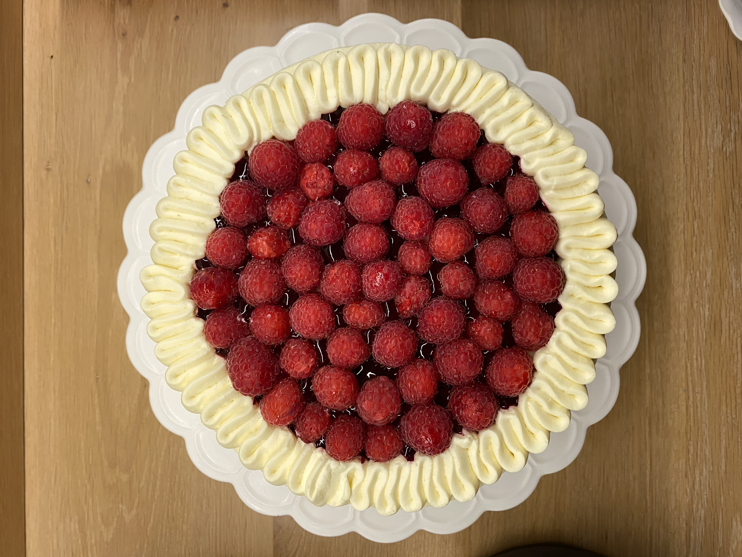 A round raspberry tart with a piped cream border on a white scalloped plate, placed on a wooden table.