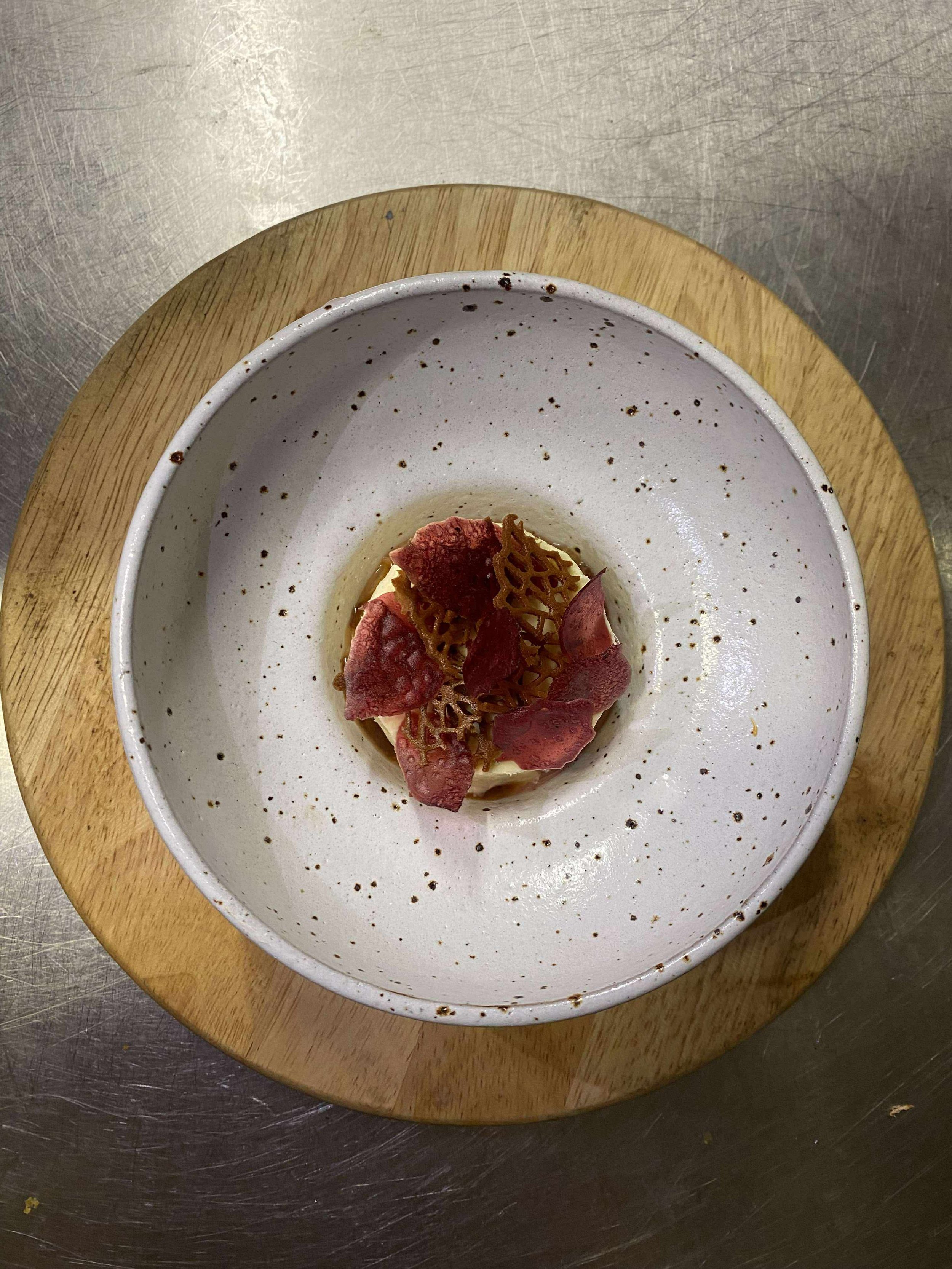A plated dessert topped with edible gold, red flower petals, and a lattice-shaped garnish, served in a speckled white bowl on a wooden plate.