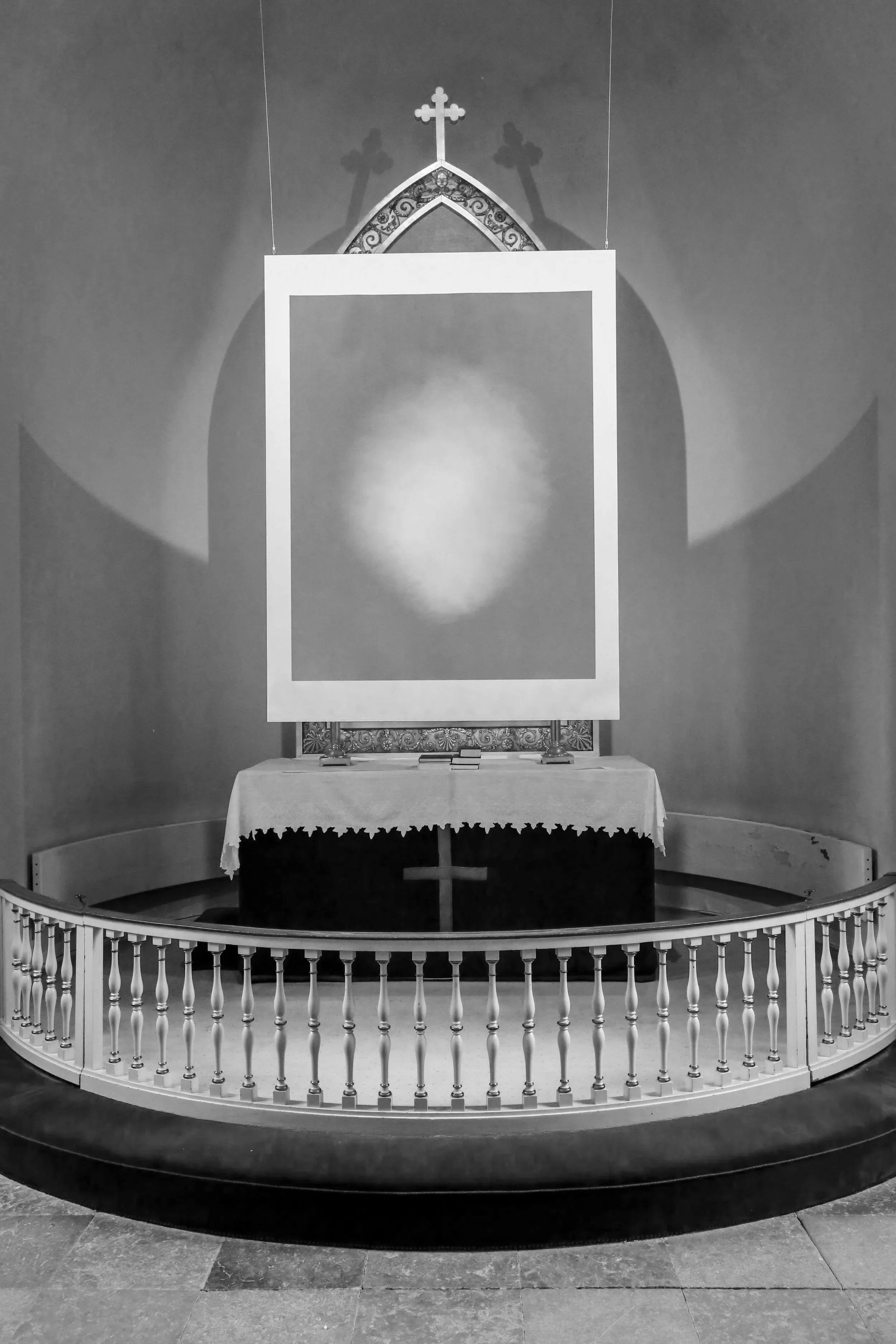 An altar with a white cloth in a church, an ornate cross and decorative wooden arch above, and a framed artwork hanging in the center.
