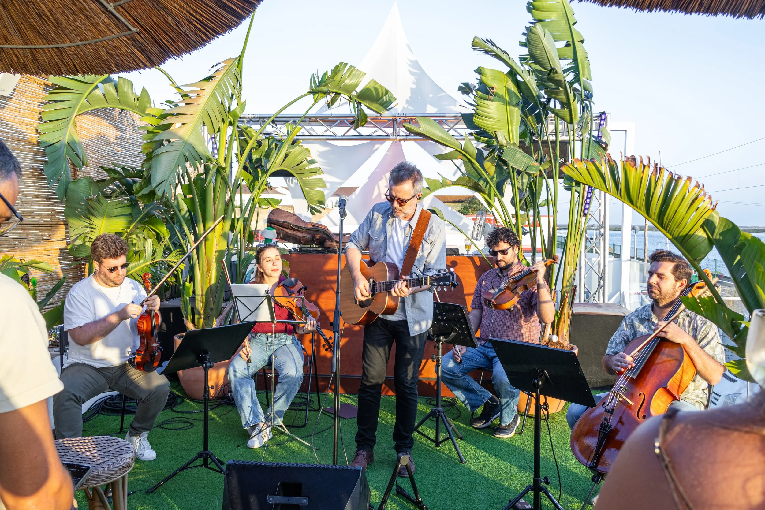 Grupo de músicos tocando violão, violino e violoncelo ao ar livre com decoração tropical e amplificação de som, durante um evento ao pôr do sol.