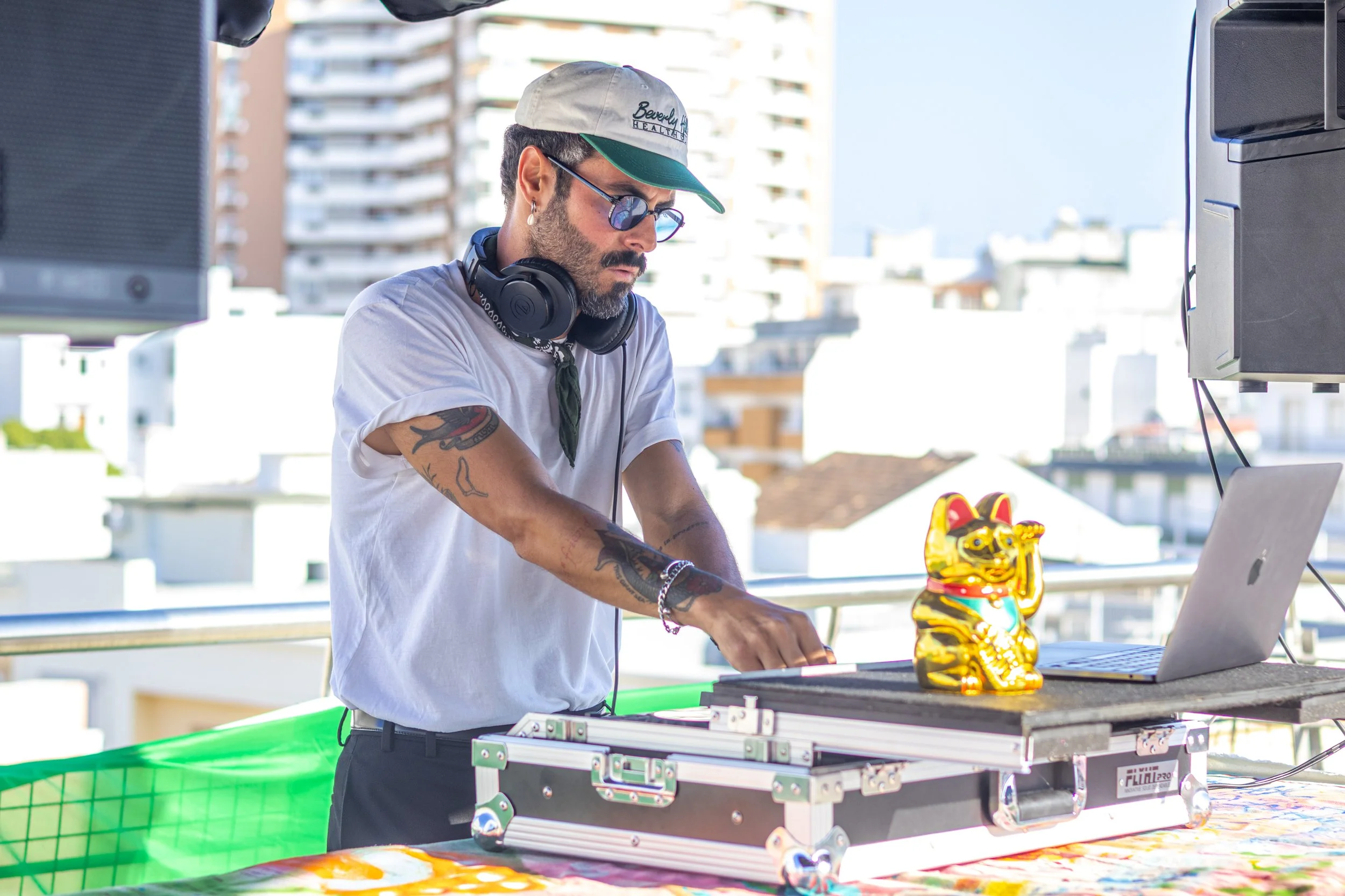 Homem DJ tocando música ao ar livre em uma cidade, usando óculos, boné e figurino casual, com equipamento de DJ e laptop na mesa.
