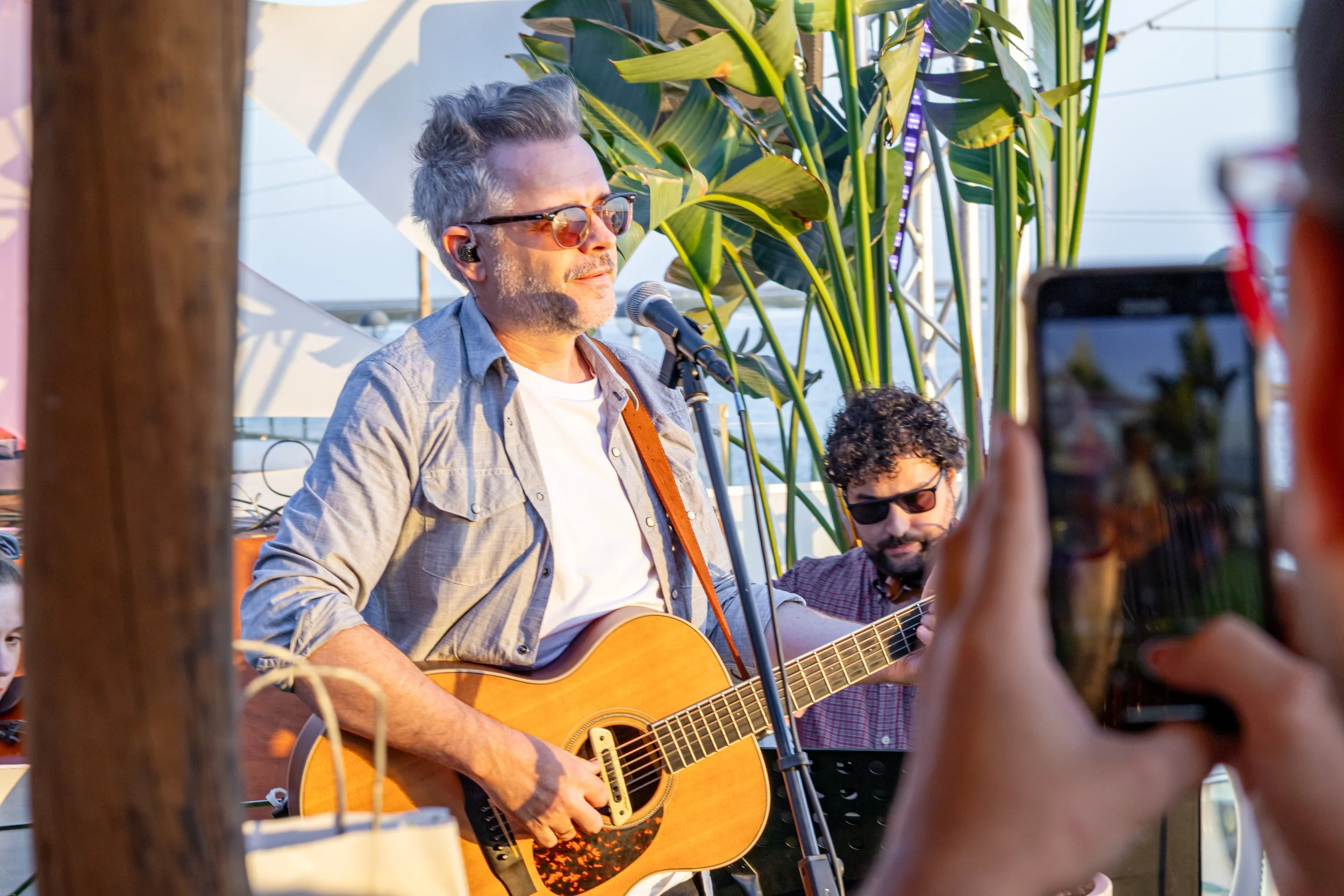 Homem tocando violão e cantando em frente a um microfone, com outros músicos ao fundo e uma pessoa tirando foto, ao ar livre com plantas verdes ao fundo.