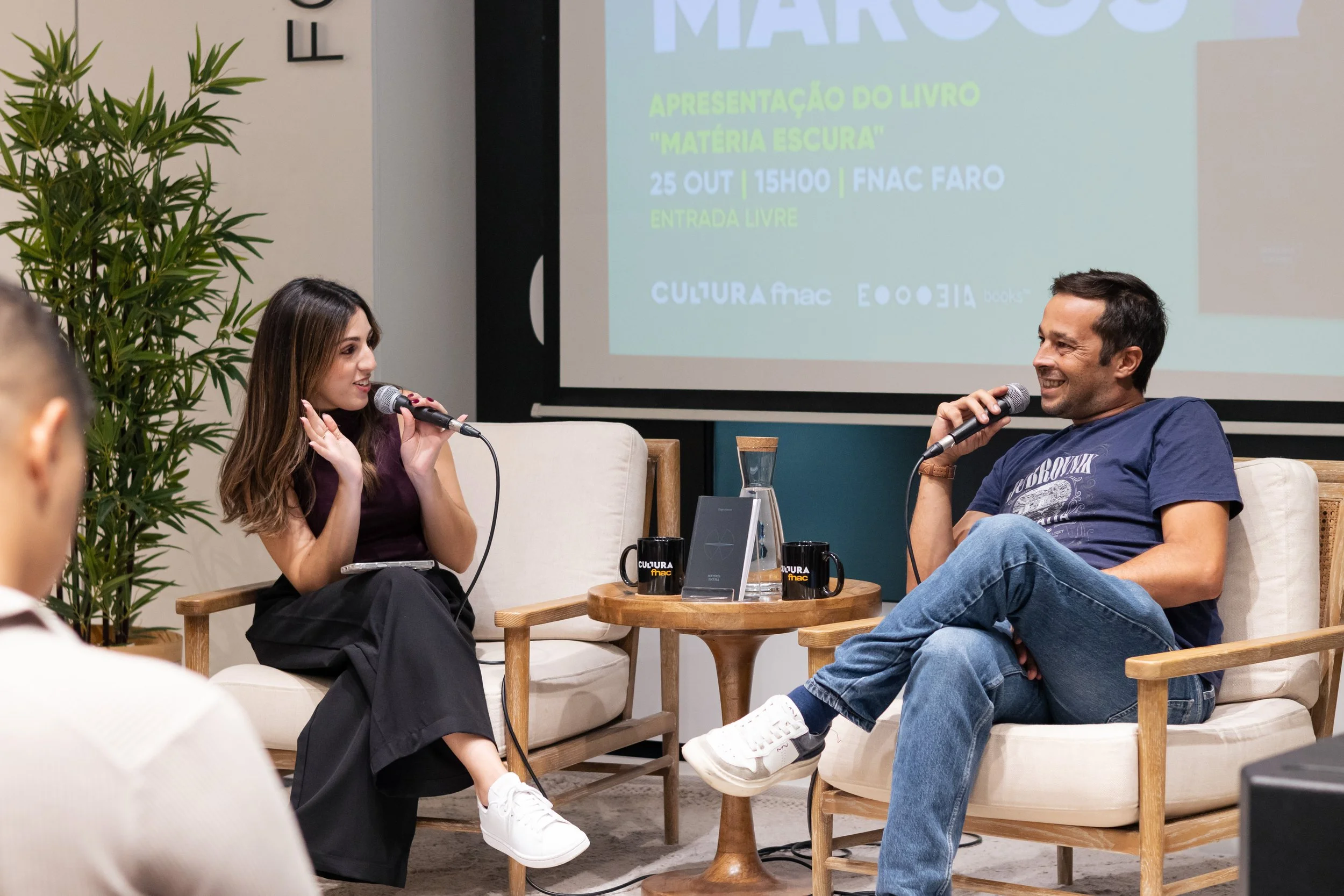 Homem e mulher conversando com microfones em cadeira de entrevista em evento na cultura FNAC, com tela de apresentação ao fundo.