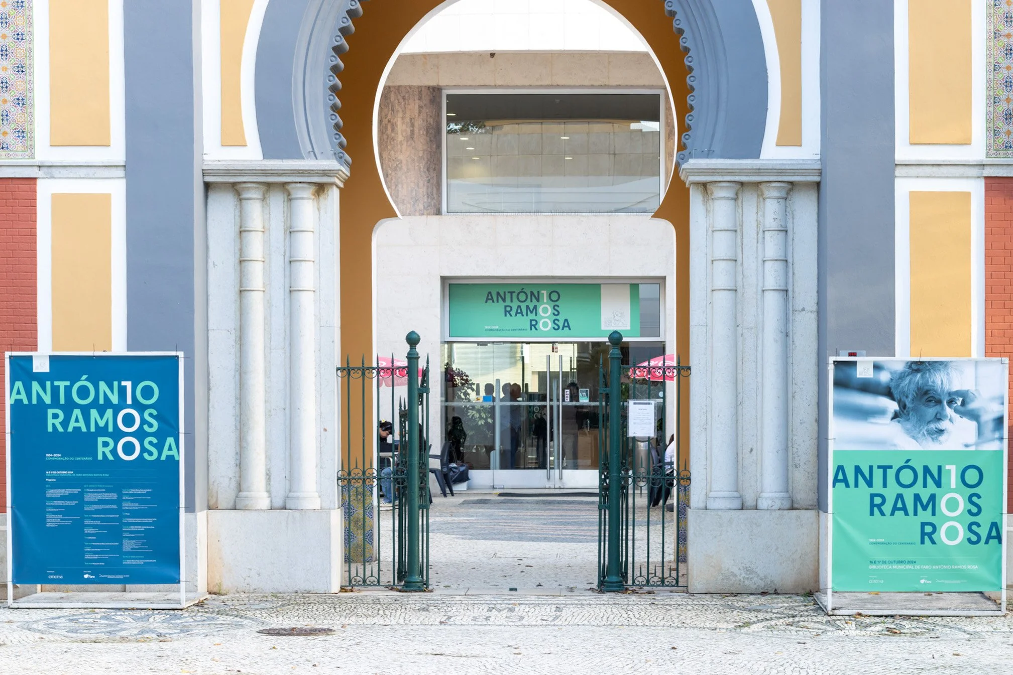 Entrada de uma biblioteca com cartazes e banners promovendo uma exposição de Antônio Ramos Rosa, com uma porta de grade de ferro e pilares de pedra, além de uma fachada colorida.