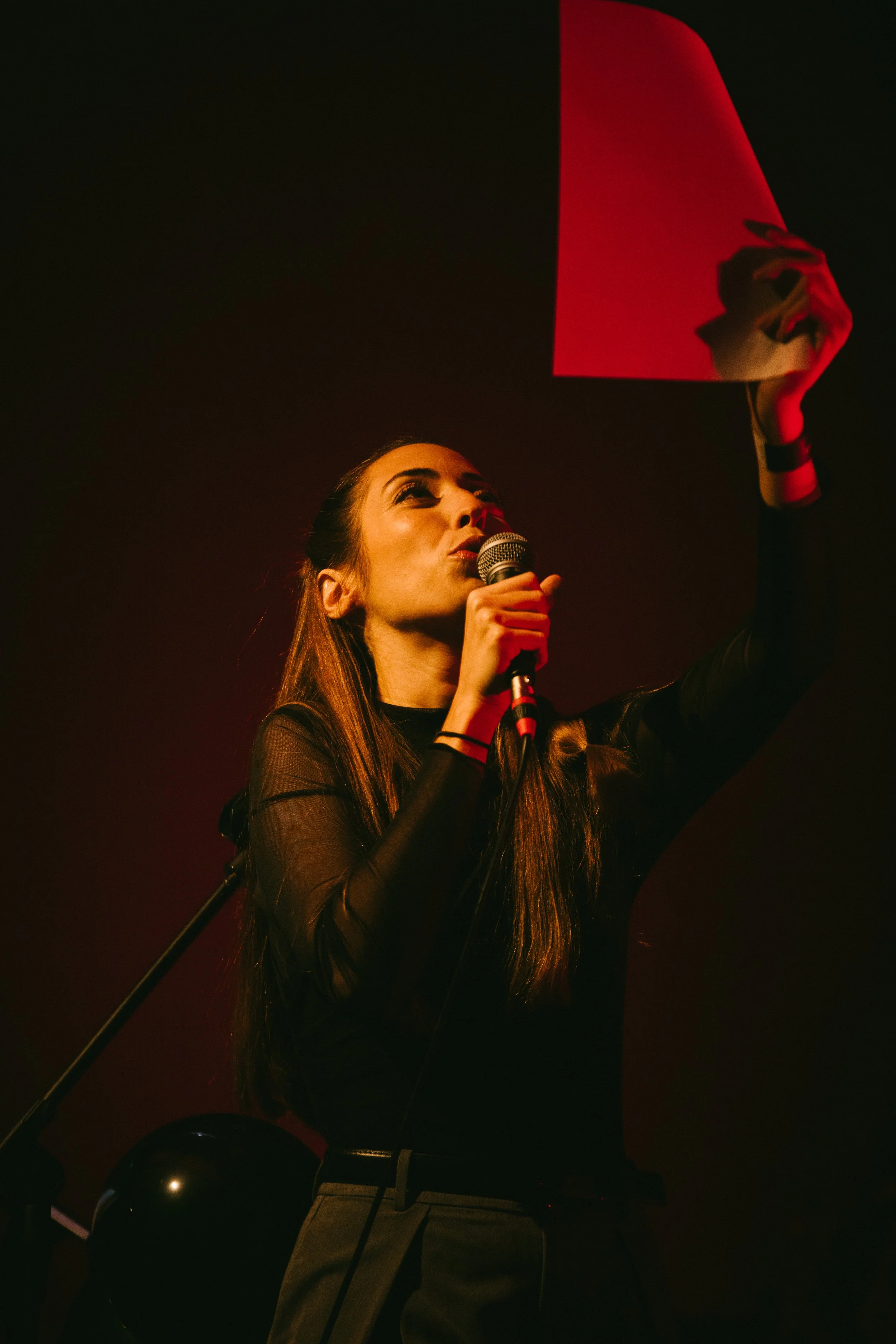 Mulher cantando no palco, segurando um microfone, com uma bandeira vermelha na mão, iluminação quente em um ambiente escuro.