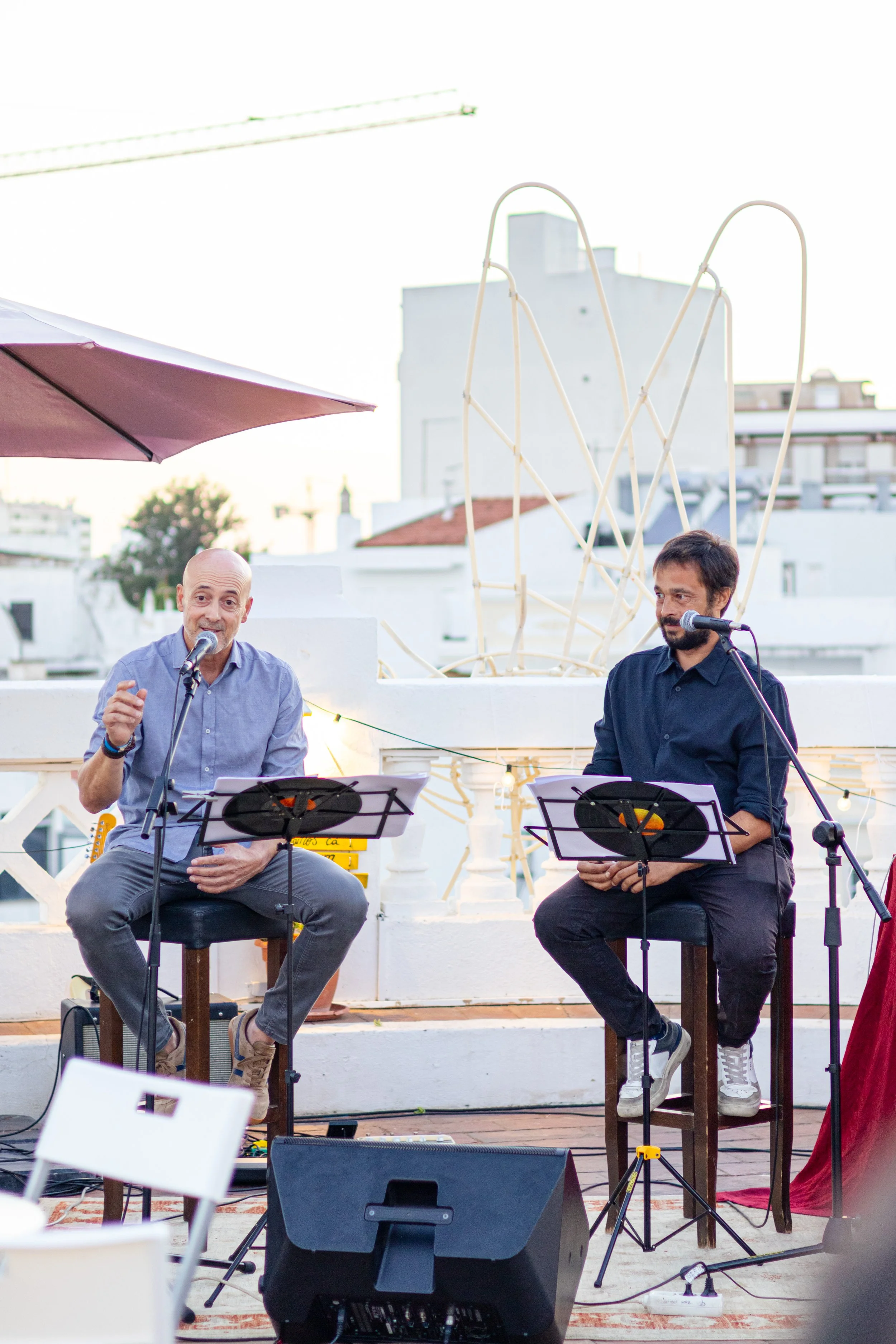 Dois homens sentados em bancos de madeira, falando em microfones durante apresentação ao ar livre, com instrumentos musicais e equipamentos ao redor, em um espaço ao ar livre com fundo de edifícios brancos e céu ao entardecer.