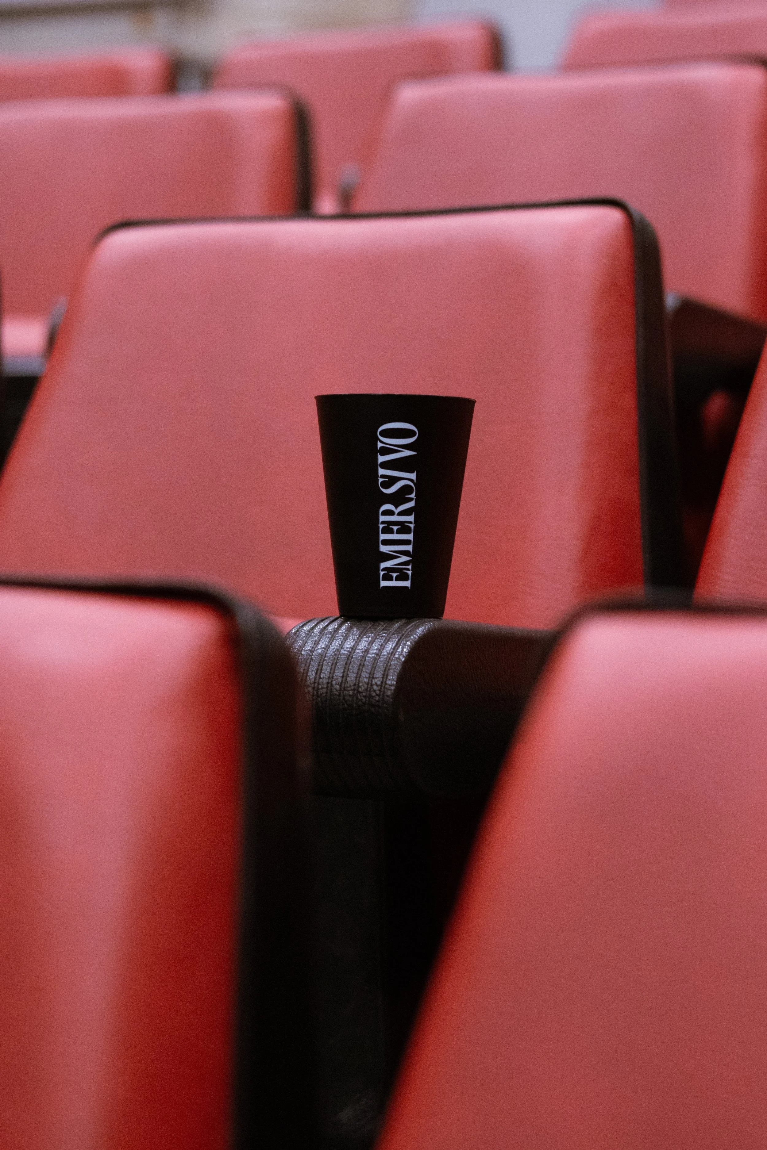 Cadeiras vermelhas de teatro ou auditório com uma garrafinha preta com a palavra "EMER VSAVO" colocada sobre uma mesa negra.