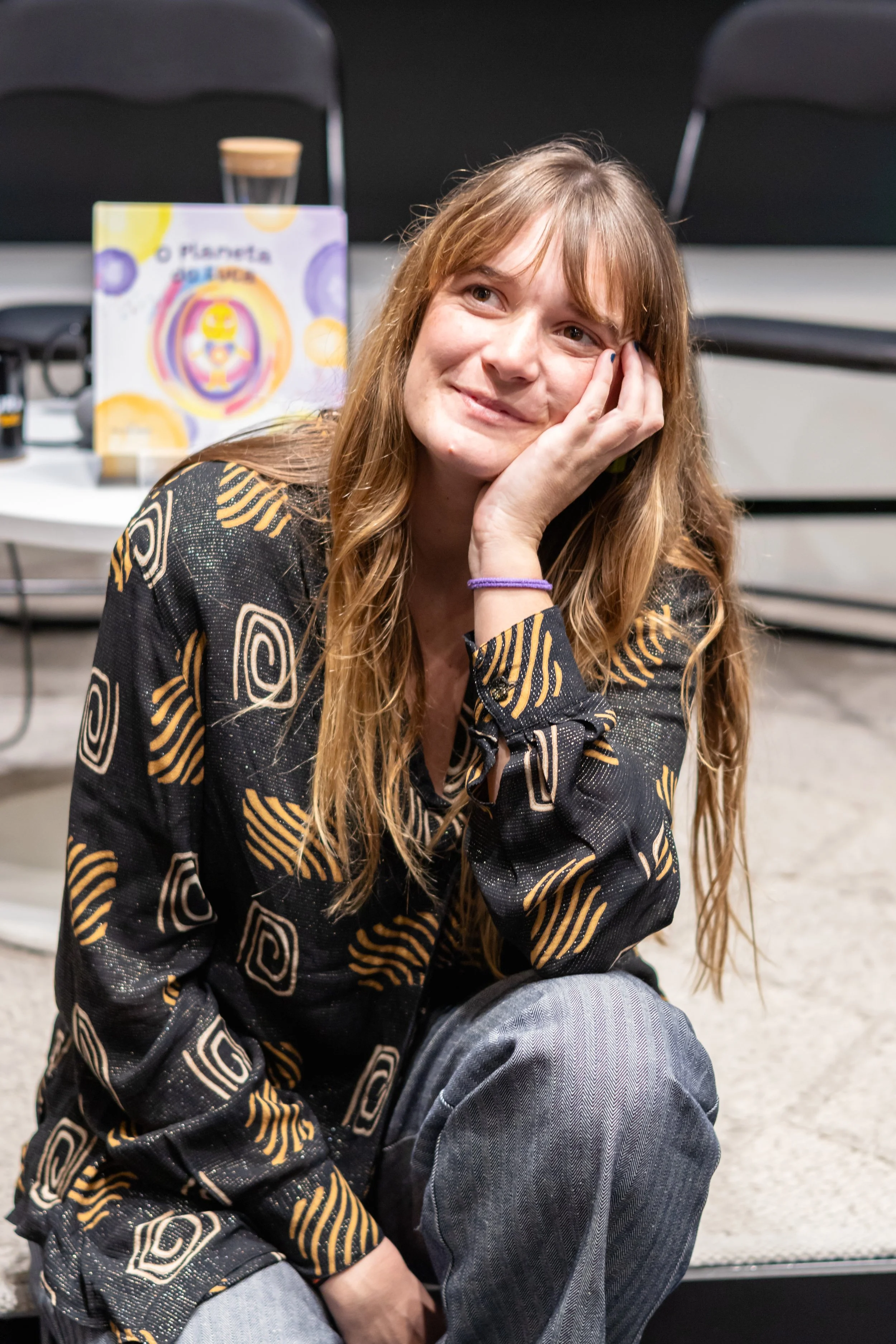 Jovem mulher de cabelo castanho claro, com roupas de estampa preta com detalhes amarelos e cinza, puxando o rosto com a mão, sorrindo levemente, sentada em piso de cimento, com assentos pretos ao fundo e livros na mesa.