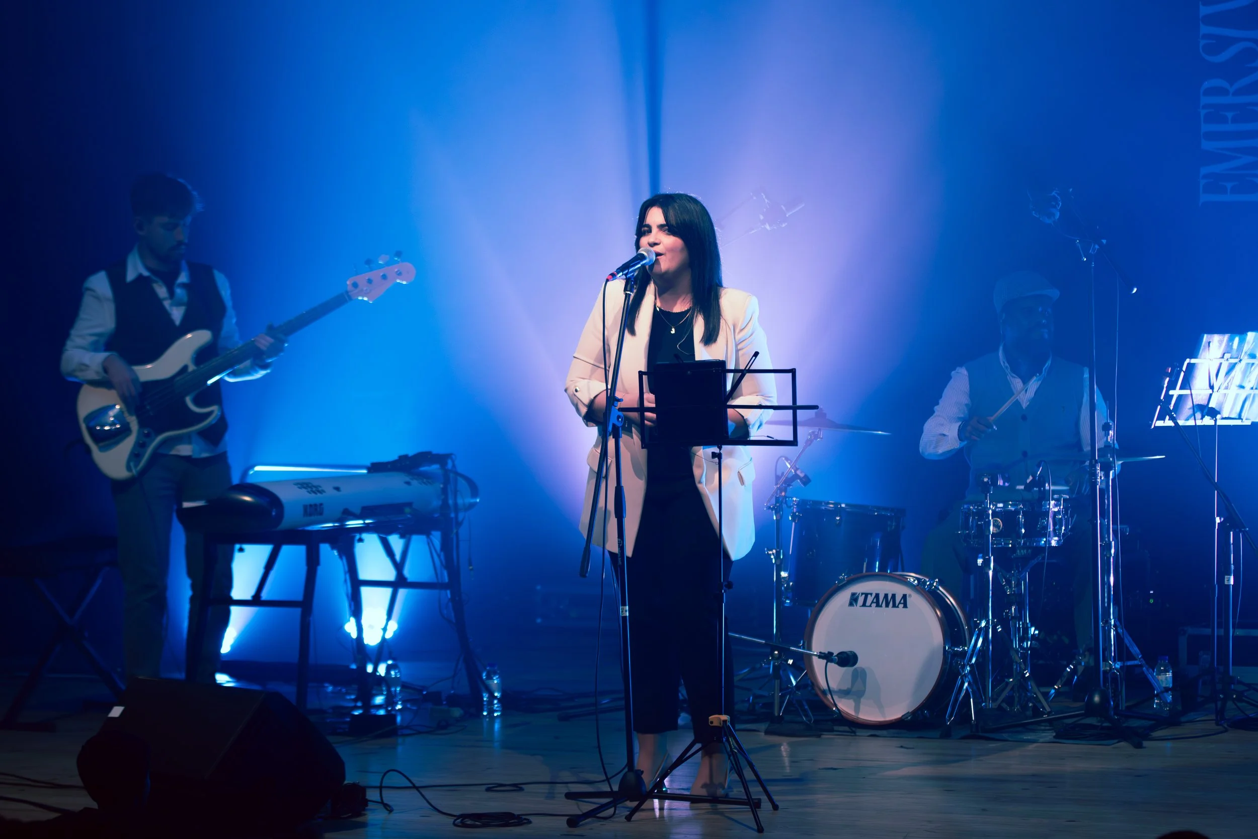 Uma mulher cantando com uma banda ao fundo em um palco com iluminação azul.