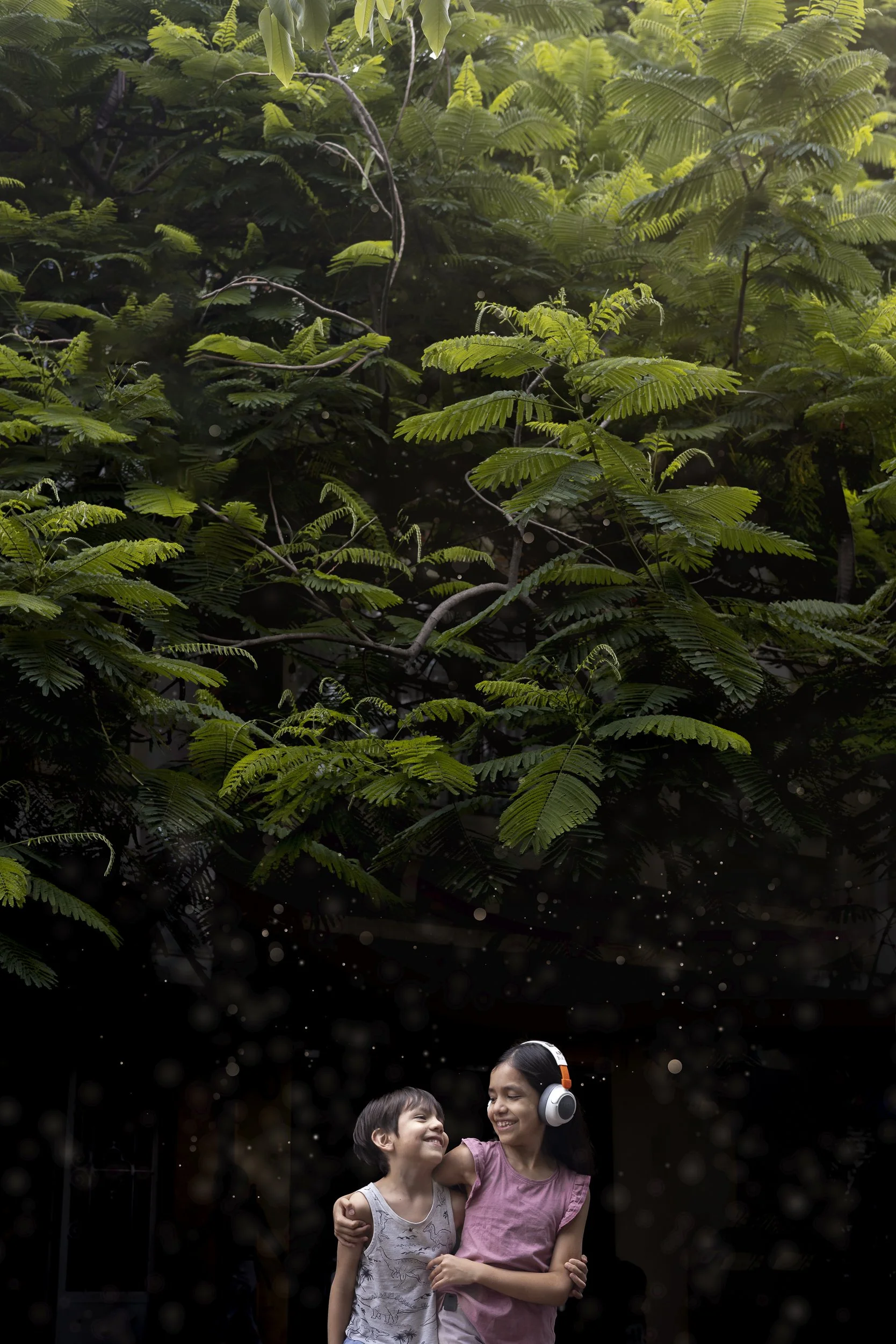 Pareja de hermanos, niña y niño abrazados, sonriendo bajo un árbol en un fondo oscuro, destacando su conexión emocional.