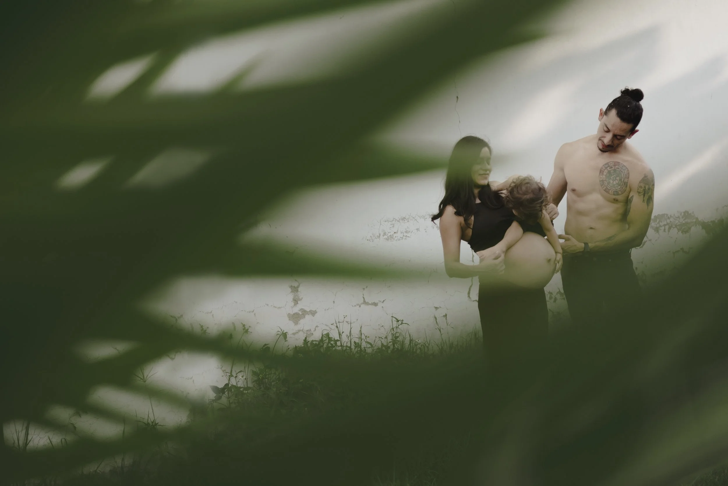 Fotografía de una familia, mujer embarazada cargando a su primer hijo y su esposo acariciándola con una sonrisa, todo enmarcado por ramas de planta desenfocadas en primer plano.