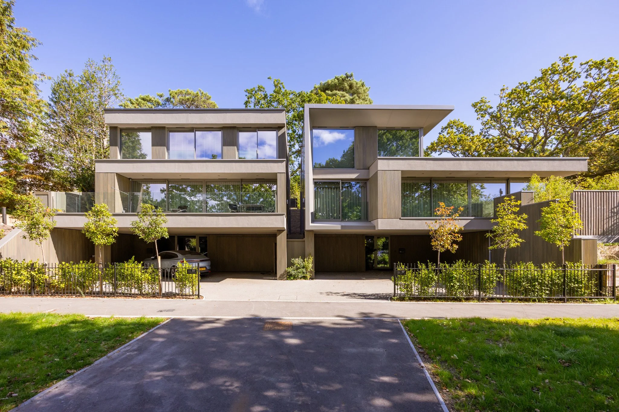 Modern two-story residential building with large glass windows and balconies, surrounded by trees and greenery, with a driveway and parking area in front.