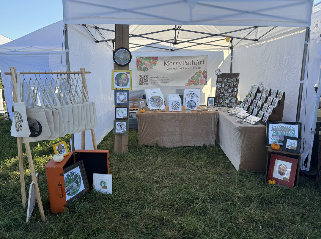 Art booth at an outdoor craft fair displaying watercolor and pen art prints, tote bags, and framed artwork under a white canopy.