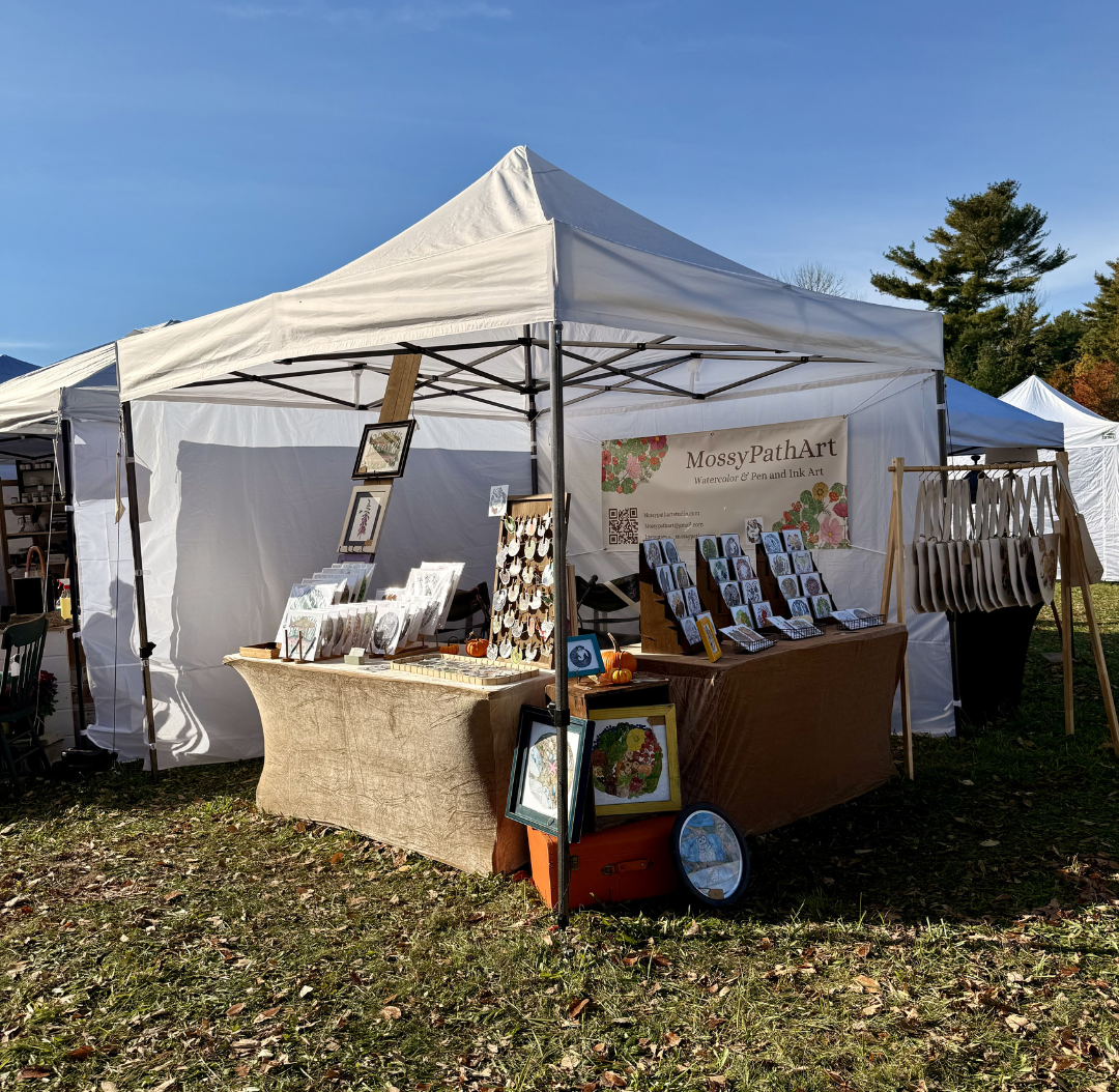 Outdoor art booth under a white canopy tent displaying watercolor and pen & ink art pieces at a fair, with trees and other tents in the background.