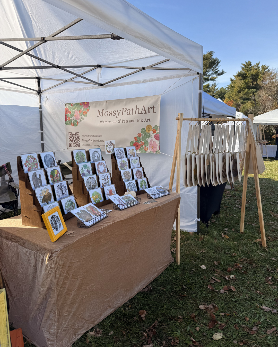 Outdoor art booth with watercolor and pen art works on display, featuring a table with postcards and a side rackBanner reads 'MossyPathArt Watercolor & Pen and Ink Art' with contact info and social media handles. Several trees and other tents are vis