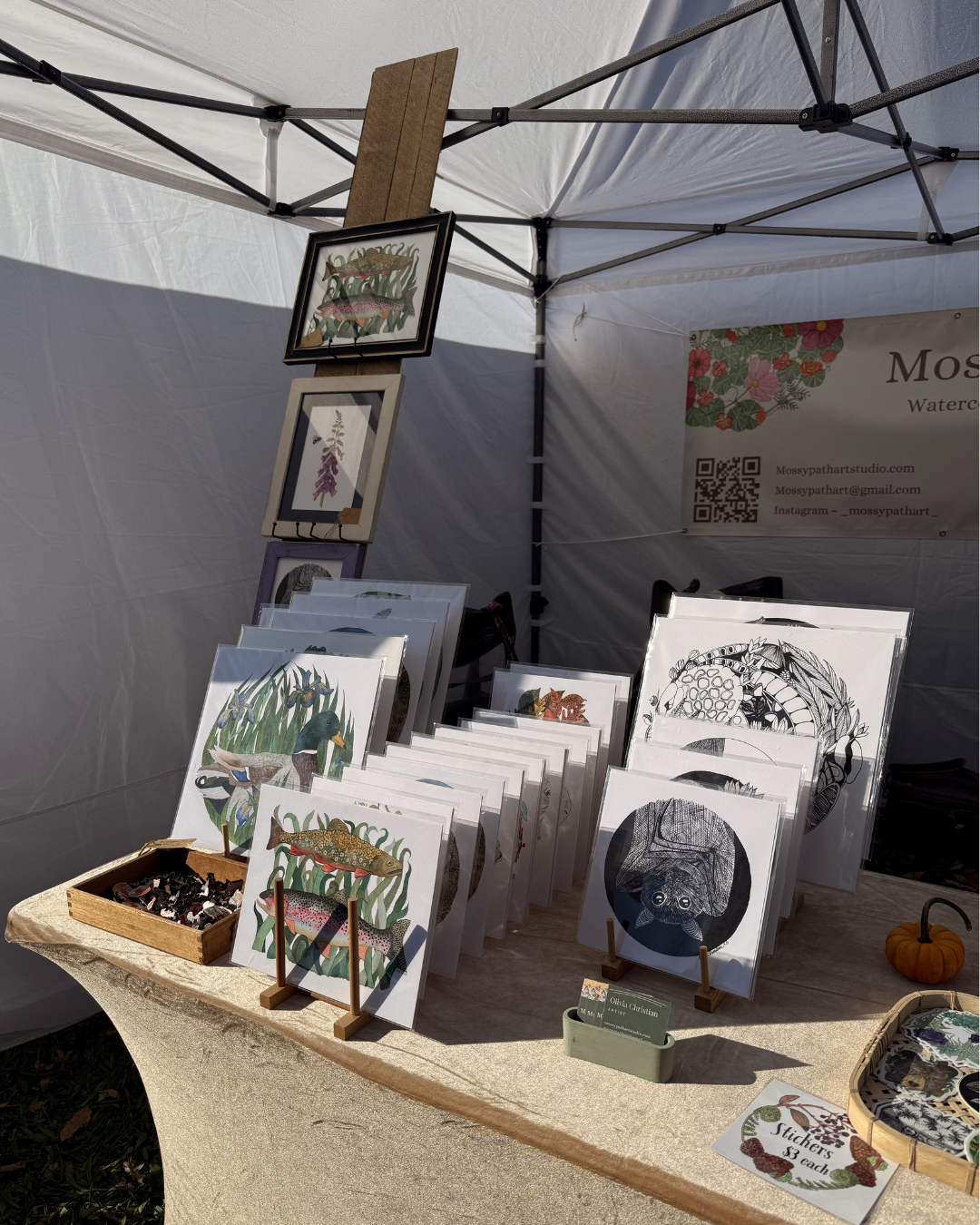Display booth showcasing various watercolor art prints of animals and nature, with printed items on a table, framed artwork hanging, and a banner with contact information.