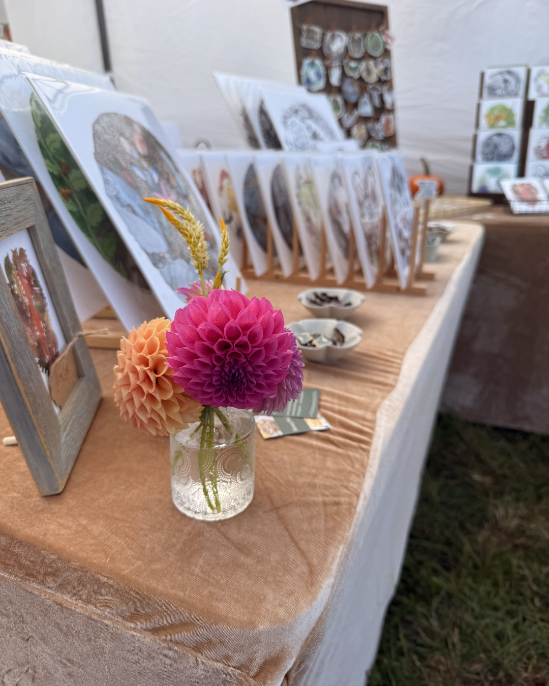 A display of artwork prints on a table with a small glass vase of pink and peach dahlias in the foreground.