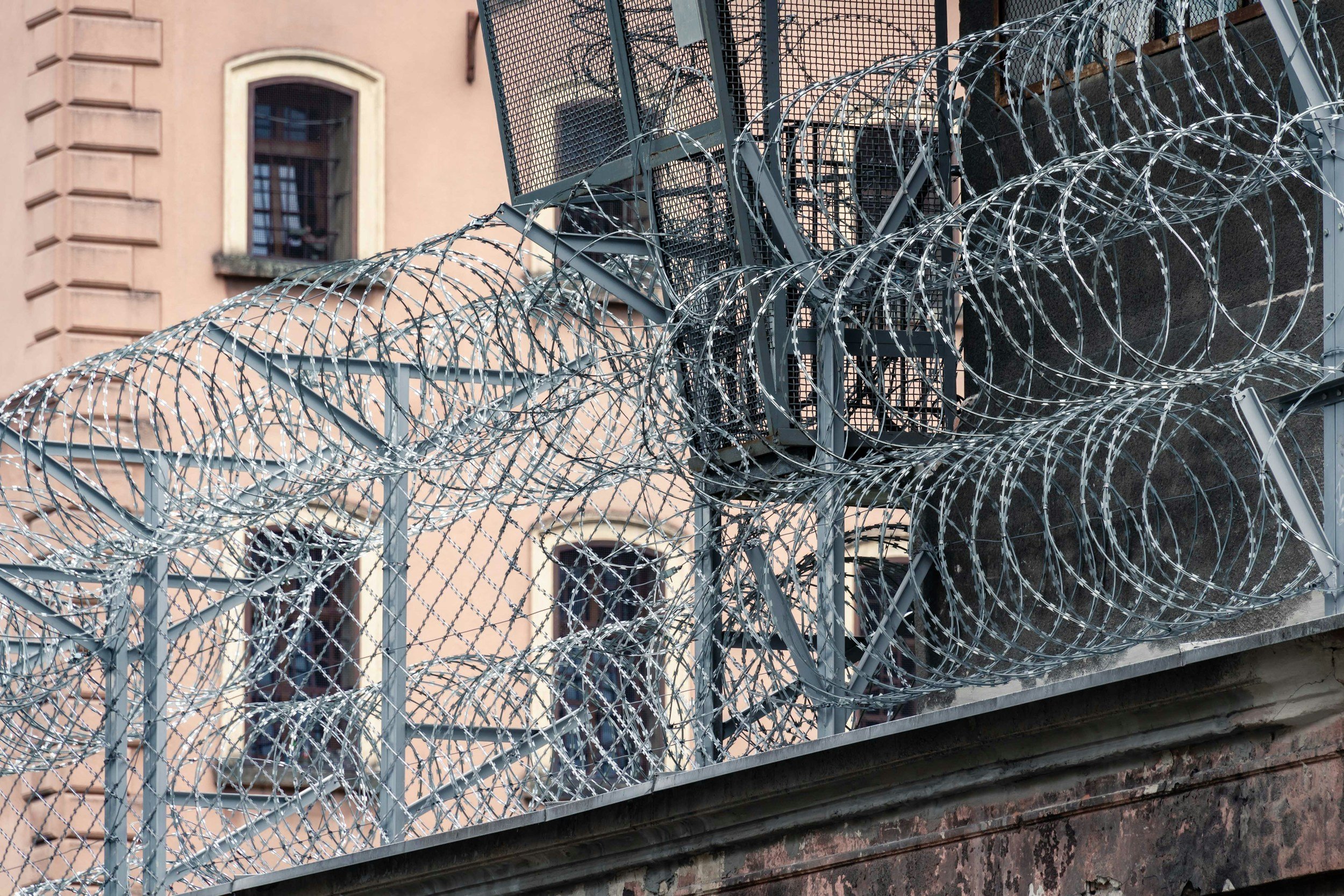 Recinzione di sicurezza in metallo con filo spinato arricciato, installata sopra un muro di mattoni, con un edificio a fianco con finestre con recinzioni.