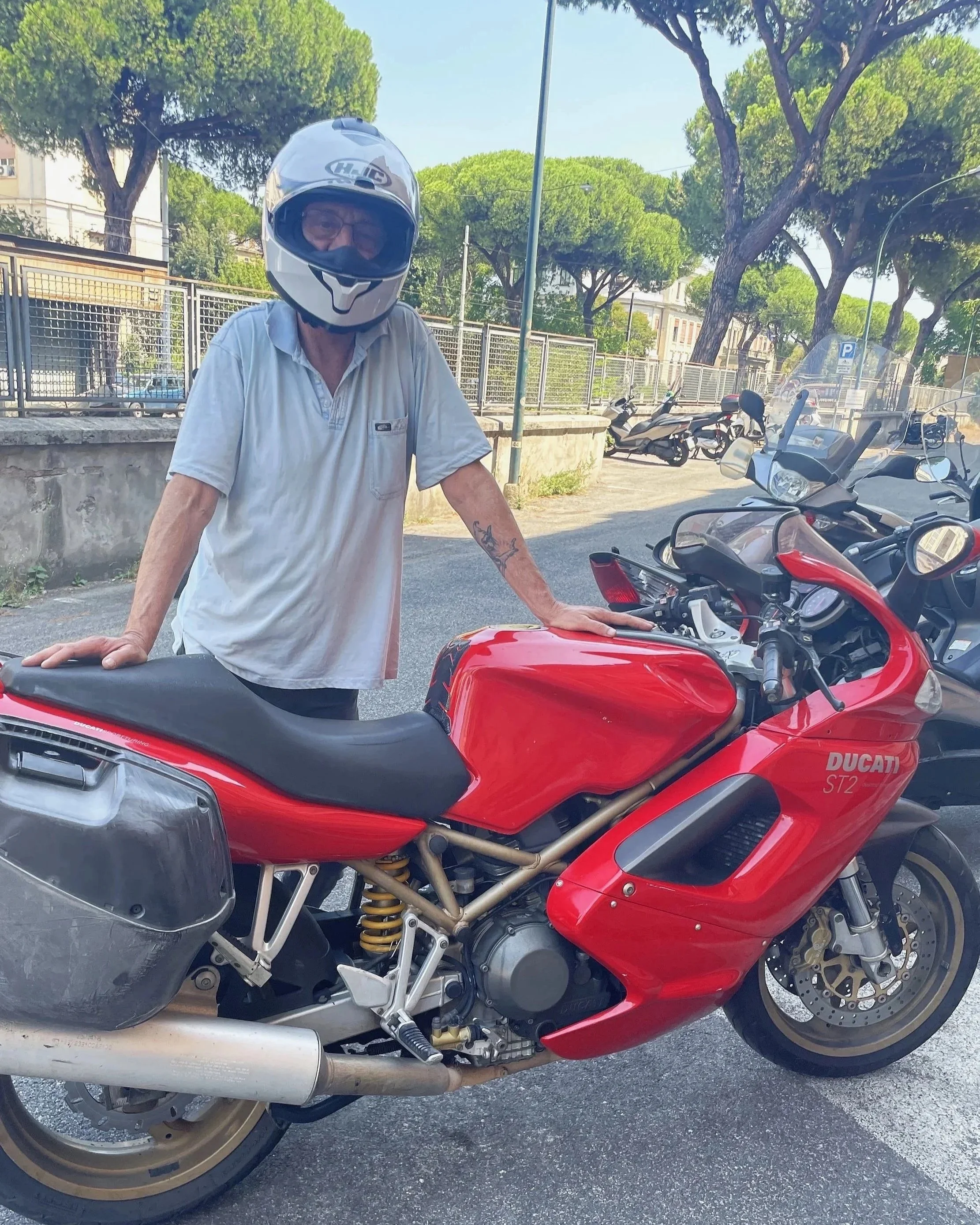Uomo con casco davanti a una motocicletta Ducati rossa in una zona con alberi e altre moto al fondo.