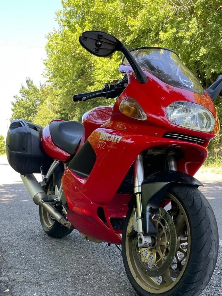 Moto Ducati Rosso con bagagli laterali nere, parcheggiata su una strada con alberi verdi sullo sfondo