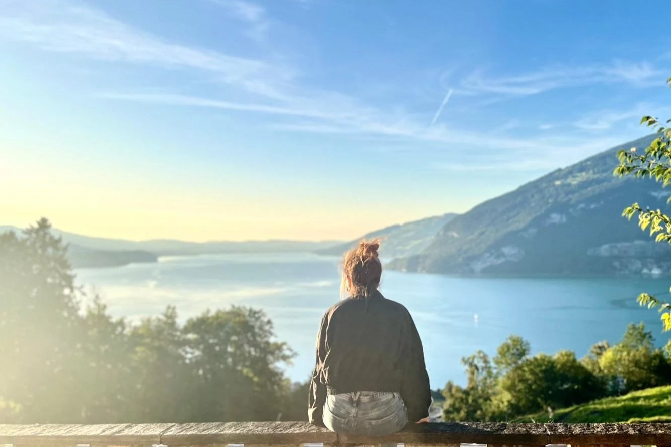 Eine Frau sitzt auf einer Holzbank und blickt auf einen See mit umliegenden Hügeln und Bergen, bei Sonnenaufgang oder Sonnenuntergang, mit blauem Himmel und Wolken.