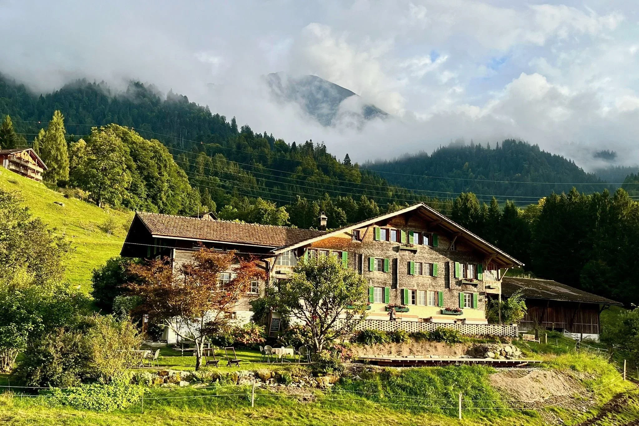 Ein Chalet-artiges Haus mit grünen Fensterläden, umgeben von Bäumen und einem Garten, auf einer Berghöhe mit Wald und Bergen im Hintergrund, teilweise von Wolken bedeckt.