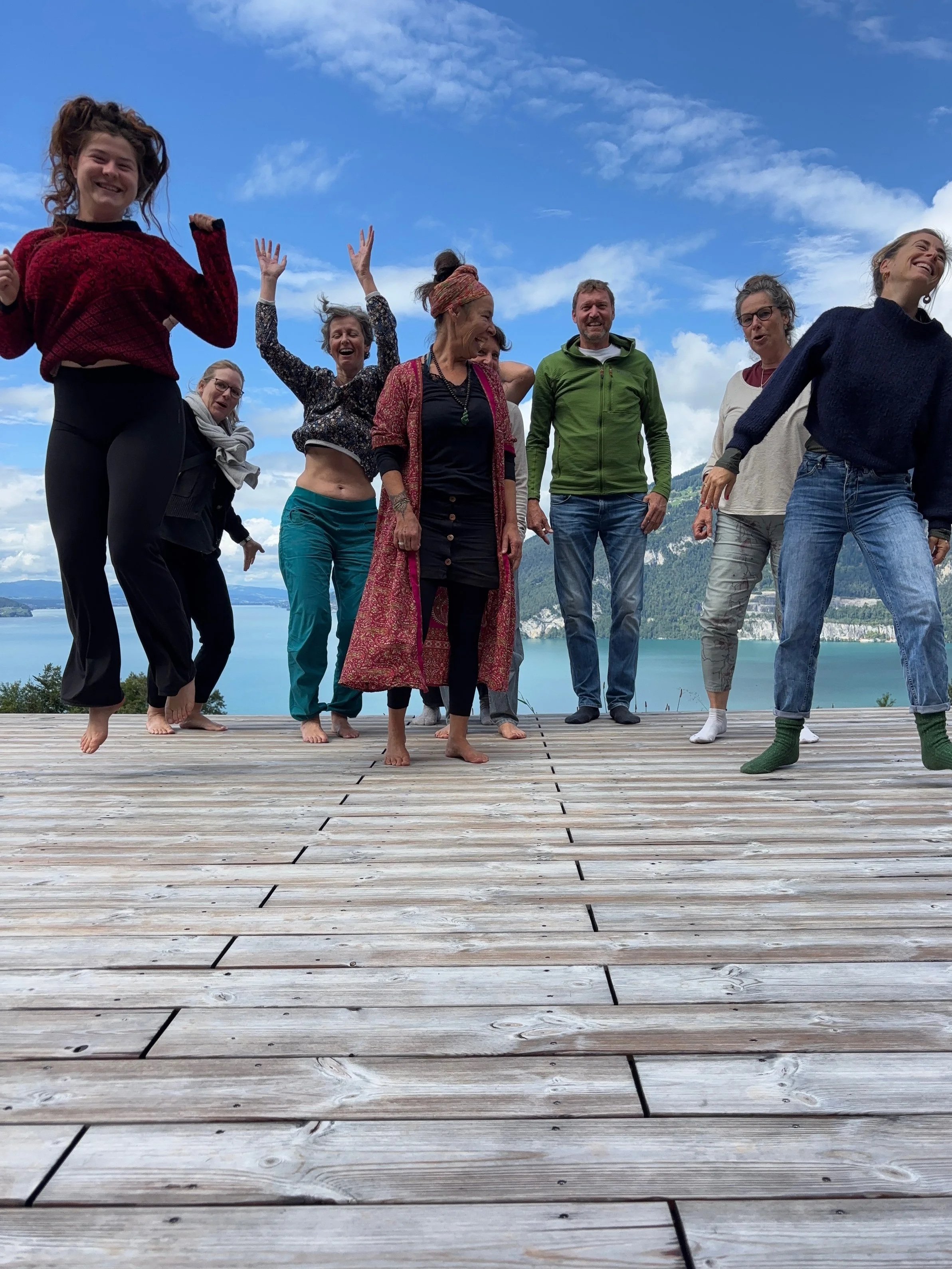 Eine Gruppe von Menschen tanzt auf einem Holzdeck mit Blick auf einen See und Berge, bei sonnigem Himmel.