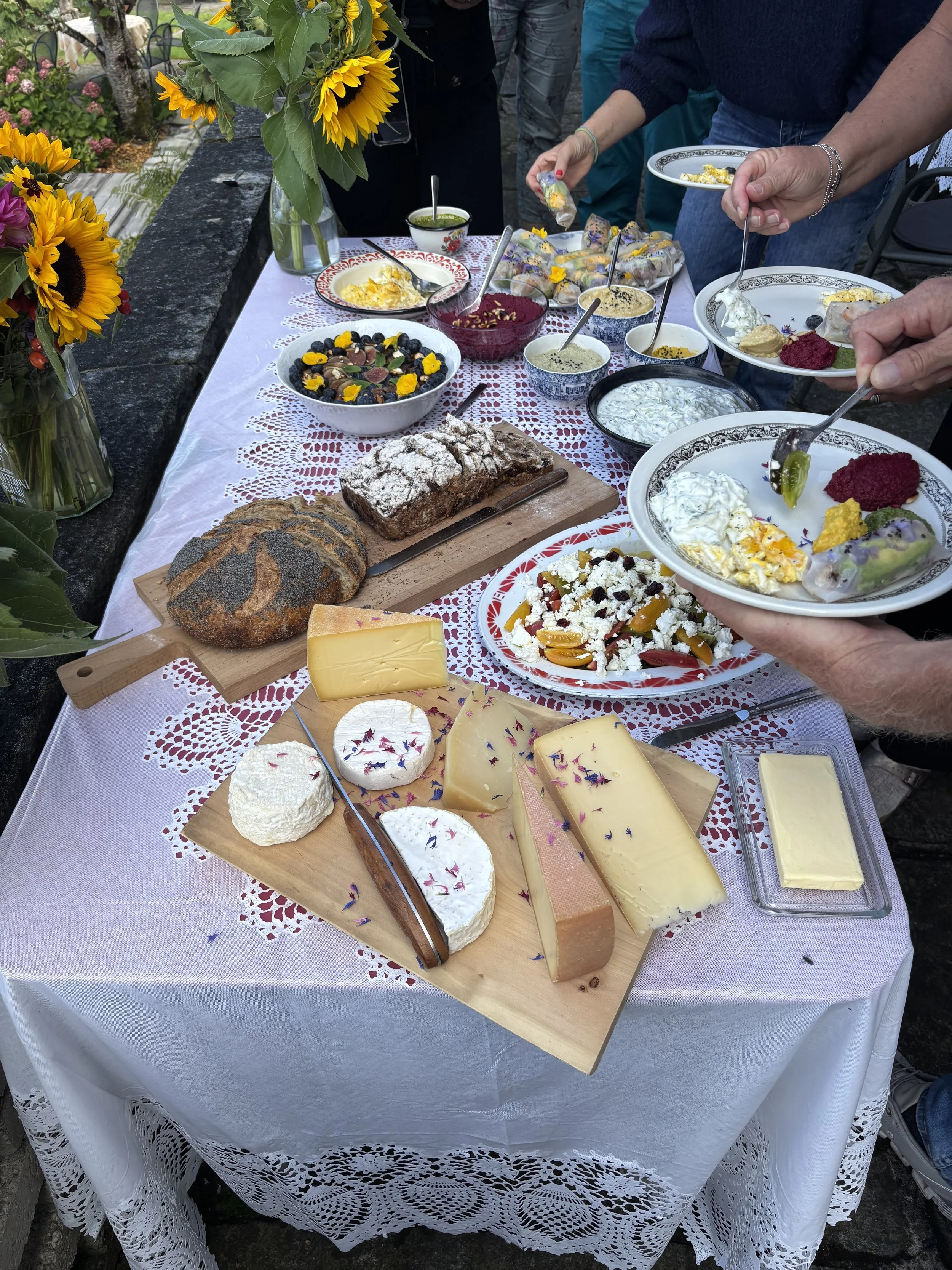 Ein festlich gedeckter Tisch mit einer Auswahl an verschiedenen Käse, frischen Blumen, Salaten, Broten, Dörrfrüchten und Suppen, der bei einer Feier oder einem Brunch genutzt wird.