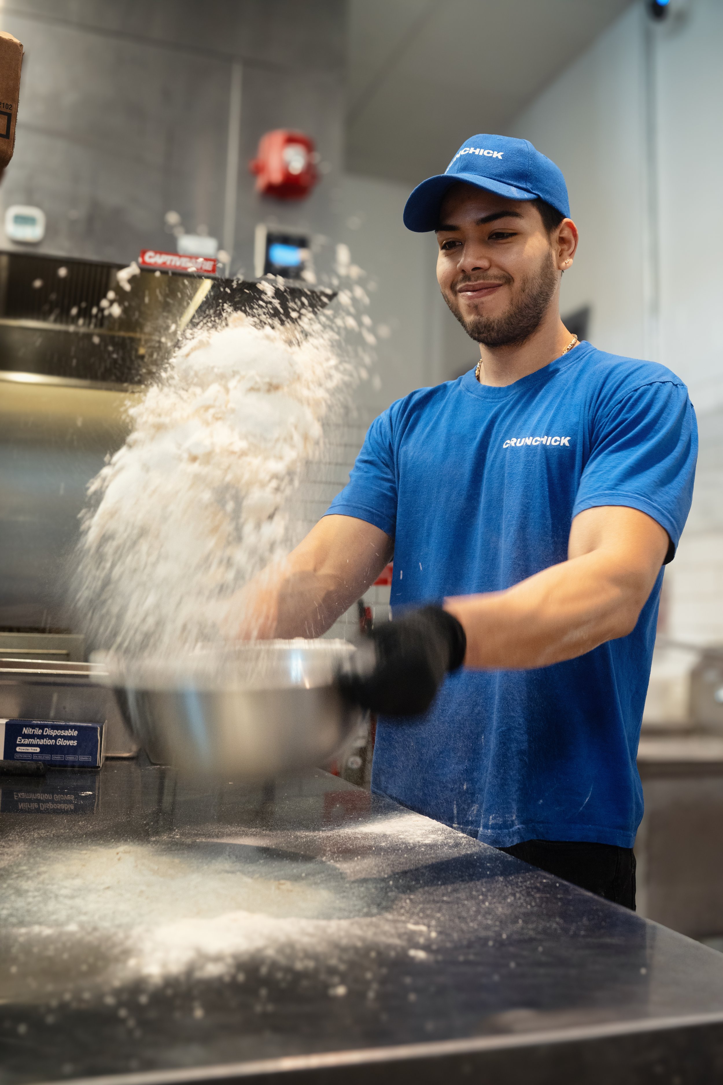 Person in blue shirt and cap preparing dough in a kitchen, tossing flour from a stainless steel bowl.