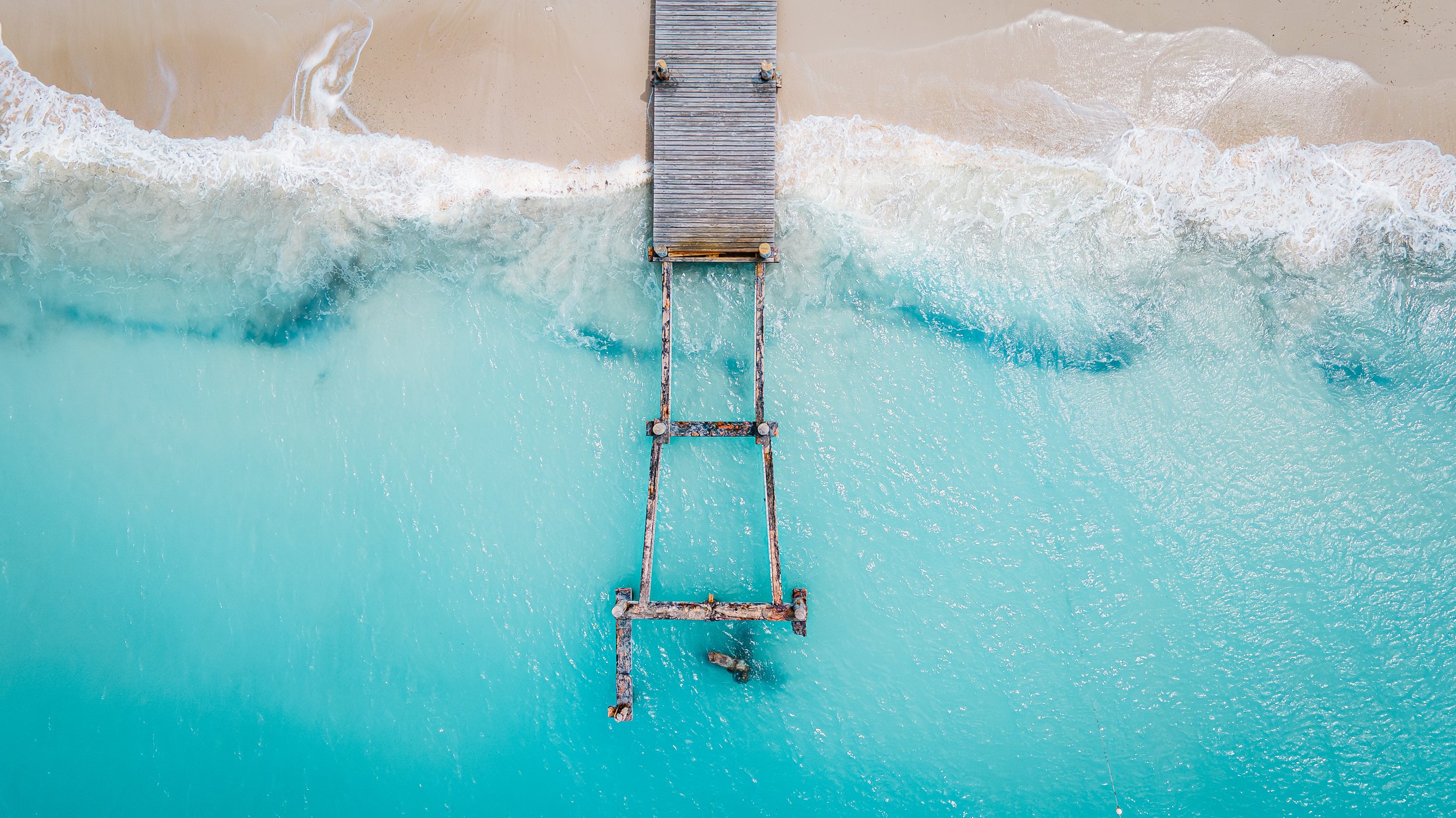 Aerial view of a wooden pier extending into turquoise ocean waves hitting a sandy beach.