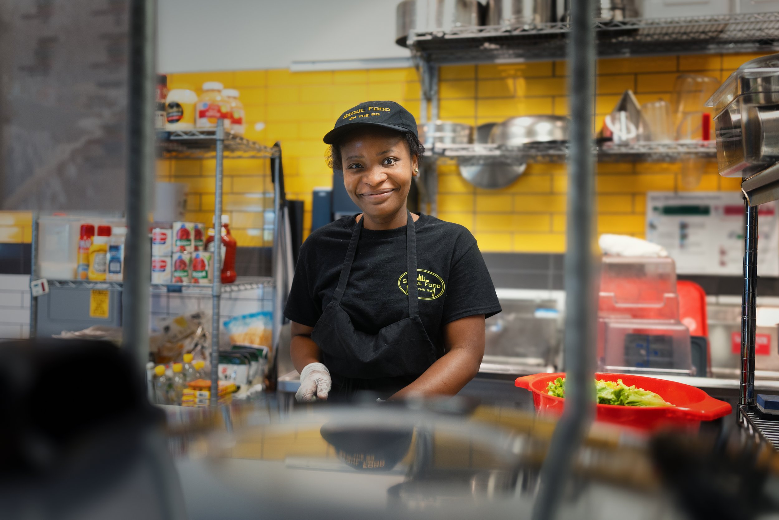 Person in a commercial kitchen wearing a black apron and hat that says 'Soul Food on the Go,' standing next to food ingredients and equipment.