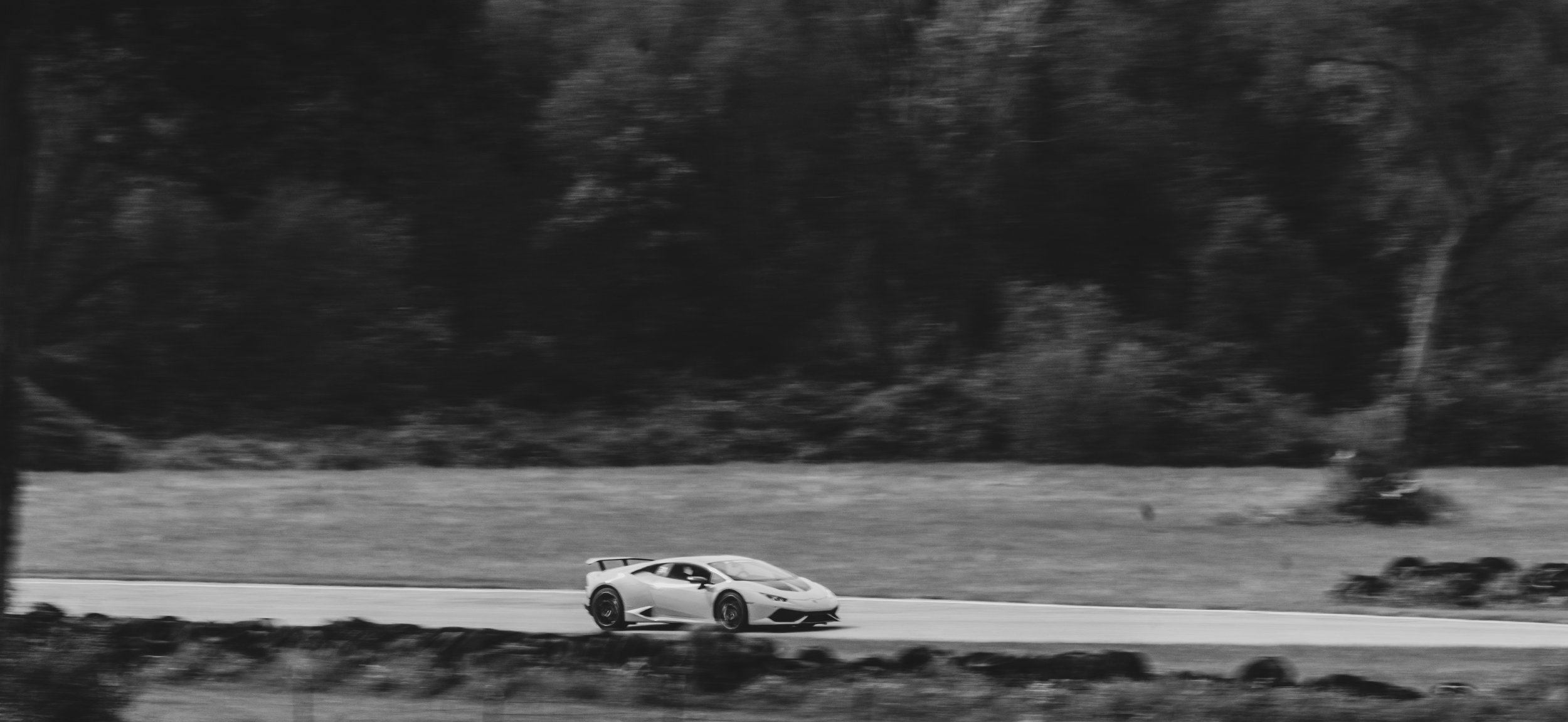 Black and white image of a sports car driving on a road with trees and grass in the background.