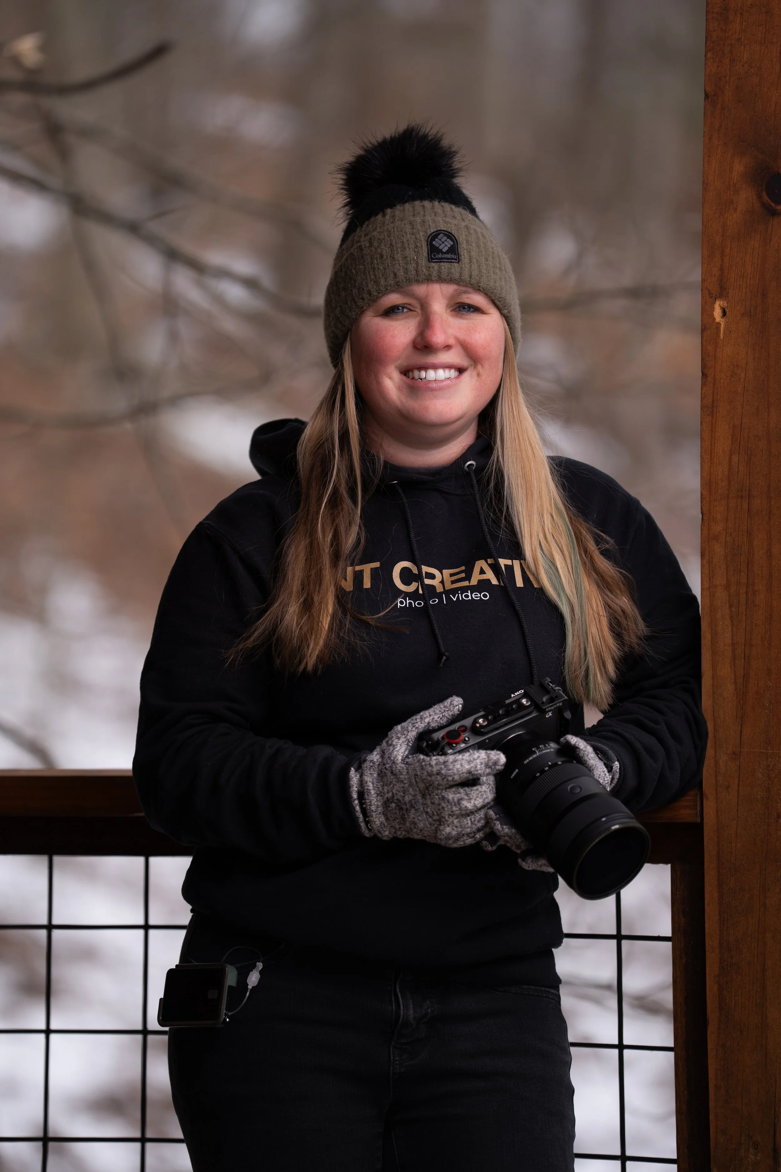 A woman posing with a camera on a deck in the winter time.