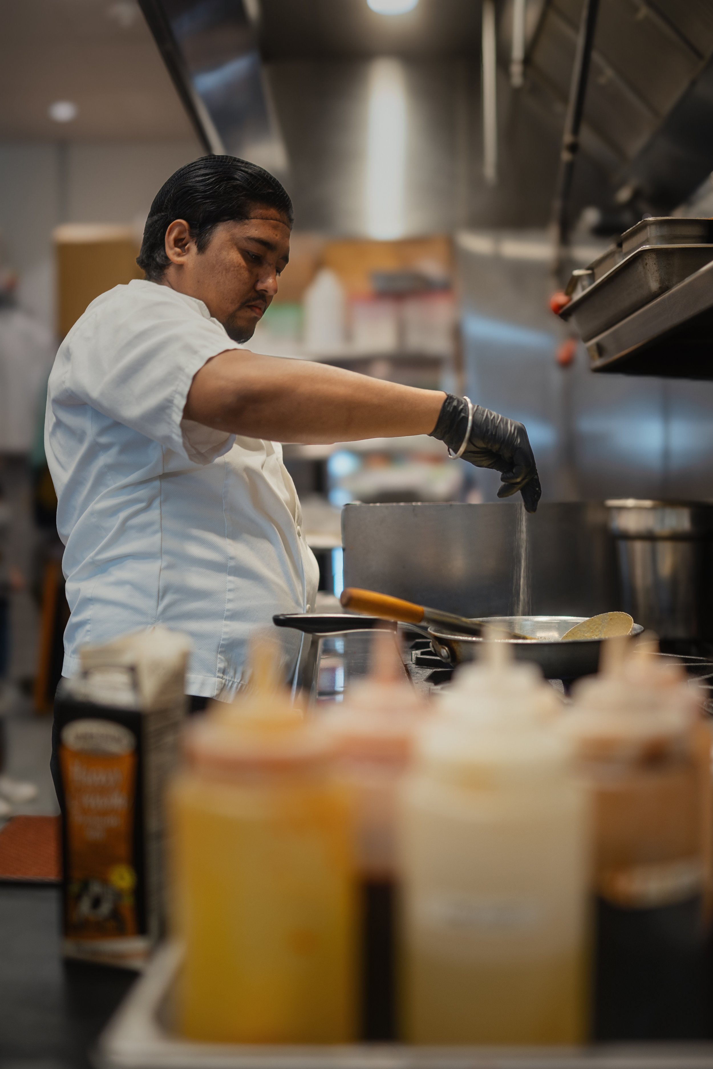 Chef seasoning food while cooking in a commercial kitchen with sauce bottles and cartons in the foreground.