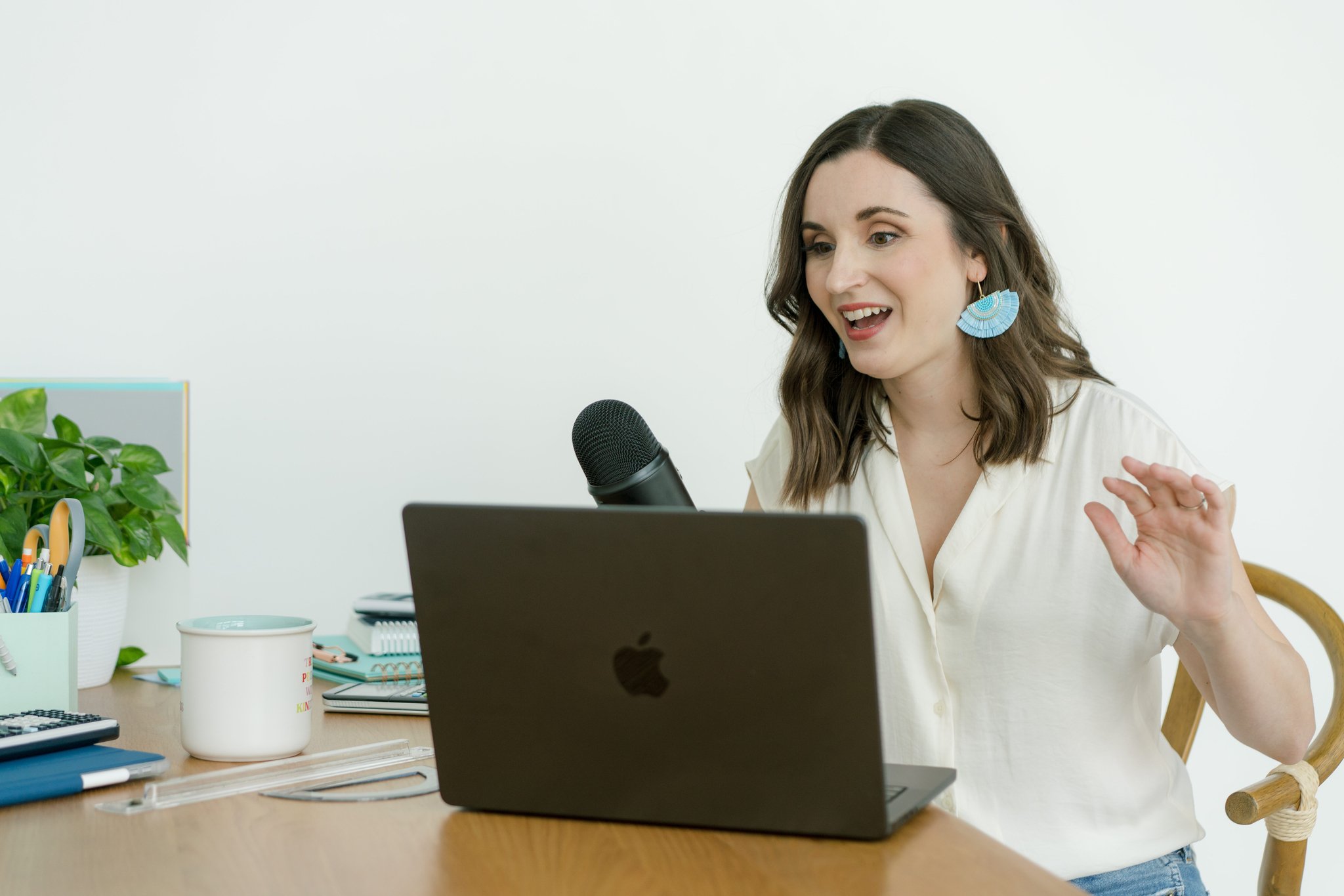 A woman with dark brown hair, wearing a white blouse and large blue earrings, is speaking into a microphone while looking at her laptop screen during a video call or recording. She is seated at a wooden desk with office supplies, a potted plant, and a white cup on it.