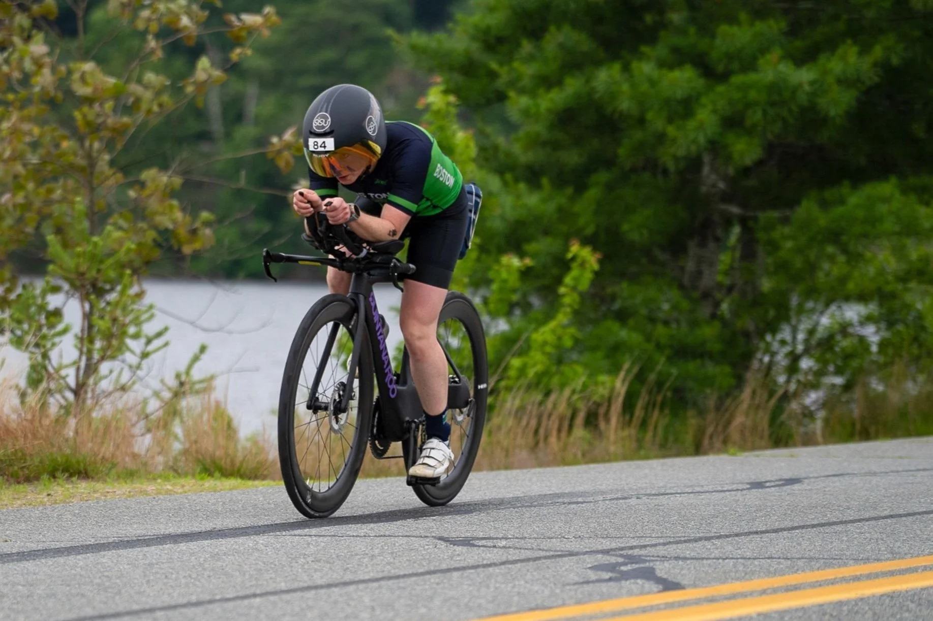 A cyclist wearing a black helmet with a white number, green and black cycling outfit, riding a black bicycle on a paved road with green trees and a body of water in the background.