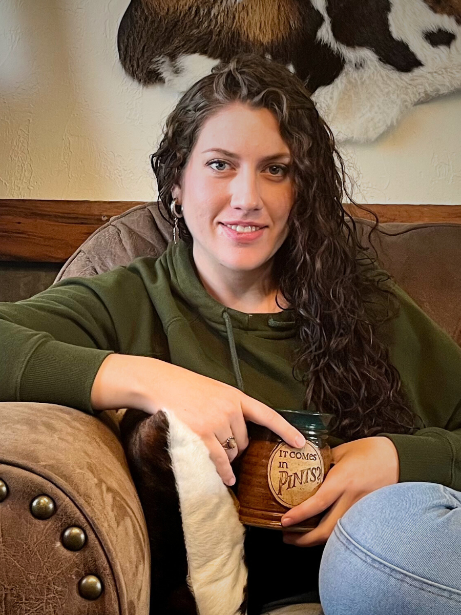 A woman with curly brown hair, wearing a green hoodie, sitting on a beige couch, holding a coffee mug