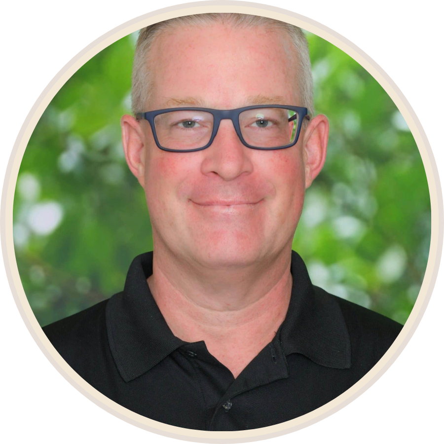 Headshot of a man with short blonde hair, glasses, and a black polo shirt, smiling in front of a green, leafy background.