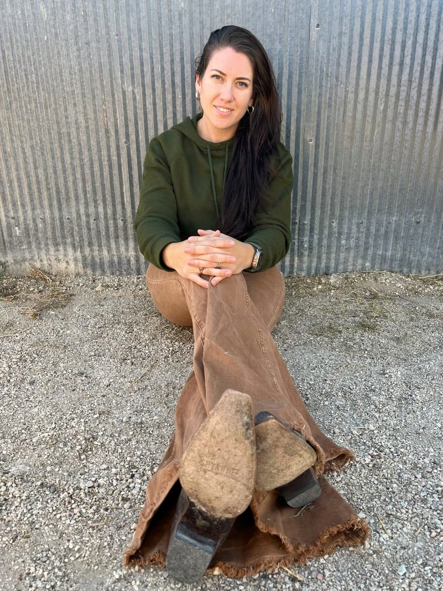 A woman with long dark hair and hoop earrings sitting outdoors against a corrugated metal wall, wearing a green hoodie and brown pants, with her feet in worn brown cowboy boots extended toward the camera.