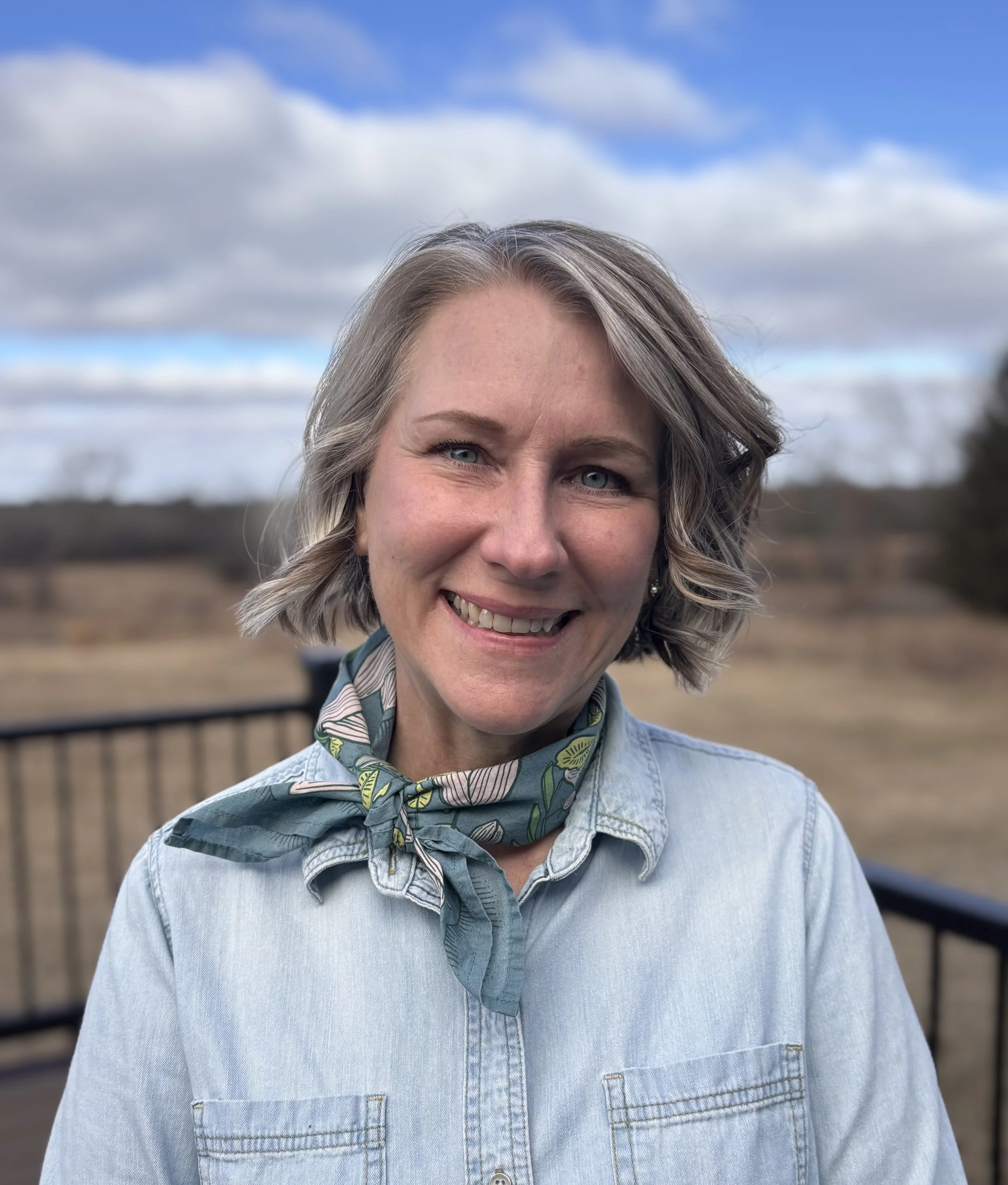 A smiling woman with short, grey hair, wearing a denim shirt and scarf, against a cloudy, grassy background