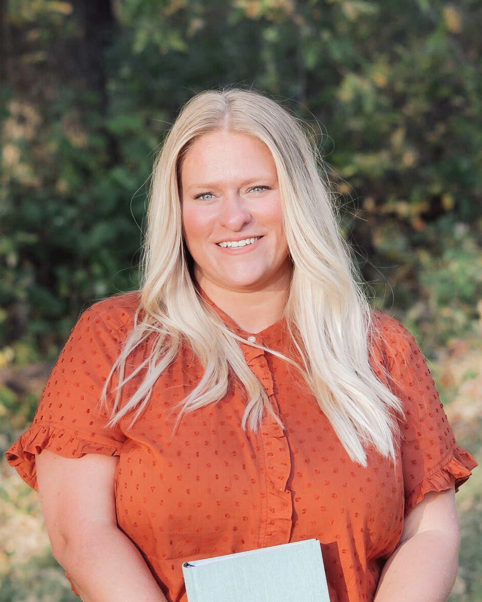 A smiling woman with long, blonde hair, wearing an orange shirt, standing outdoors with greenery in the background.