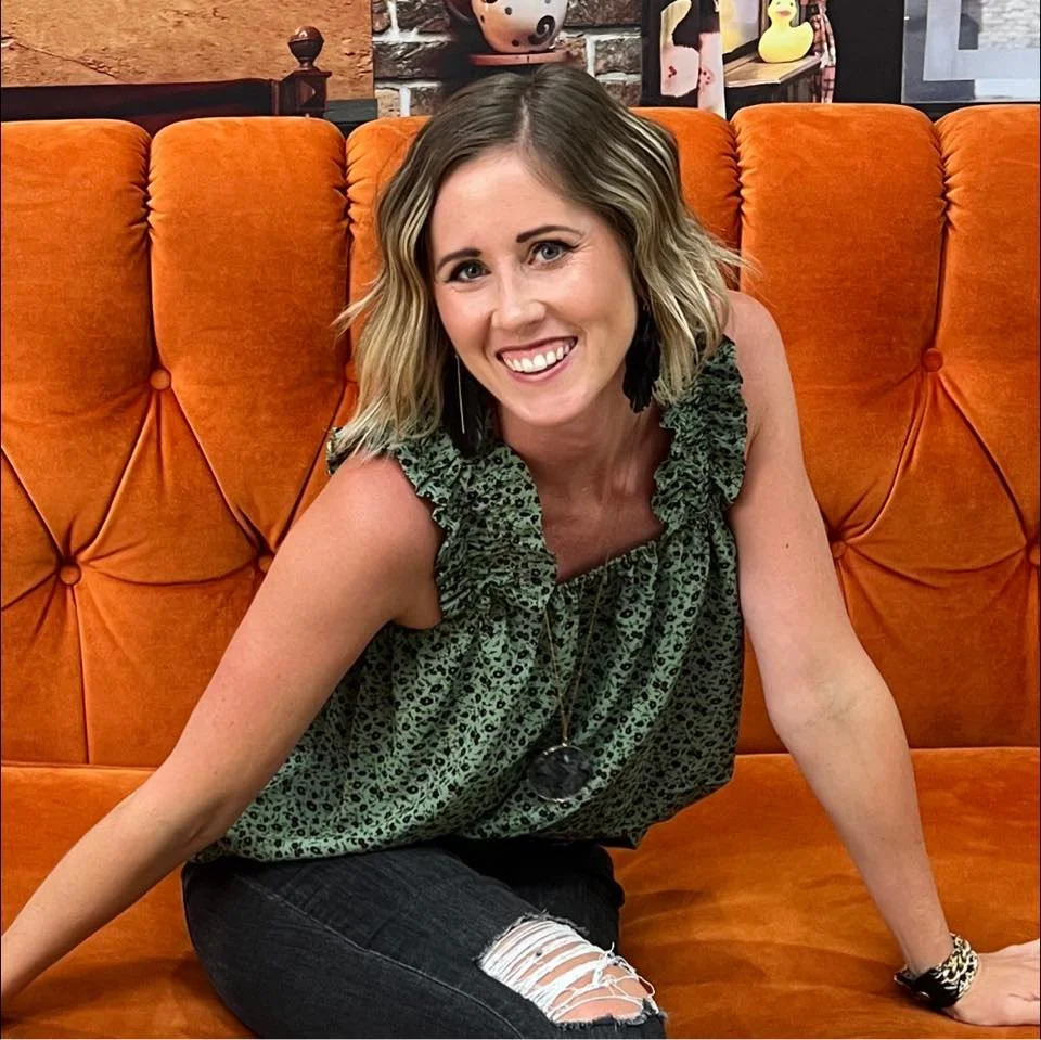 Woman with shoulder-length blonde hair, wearing a green patterned sleeveless top, ripped black jeans, and a watch, sitting on an orange tufted sofa, smiling at the camera.