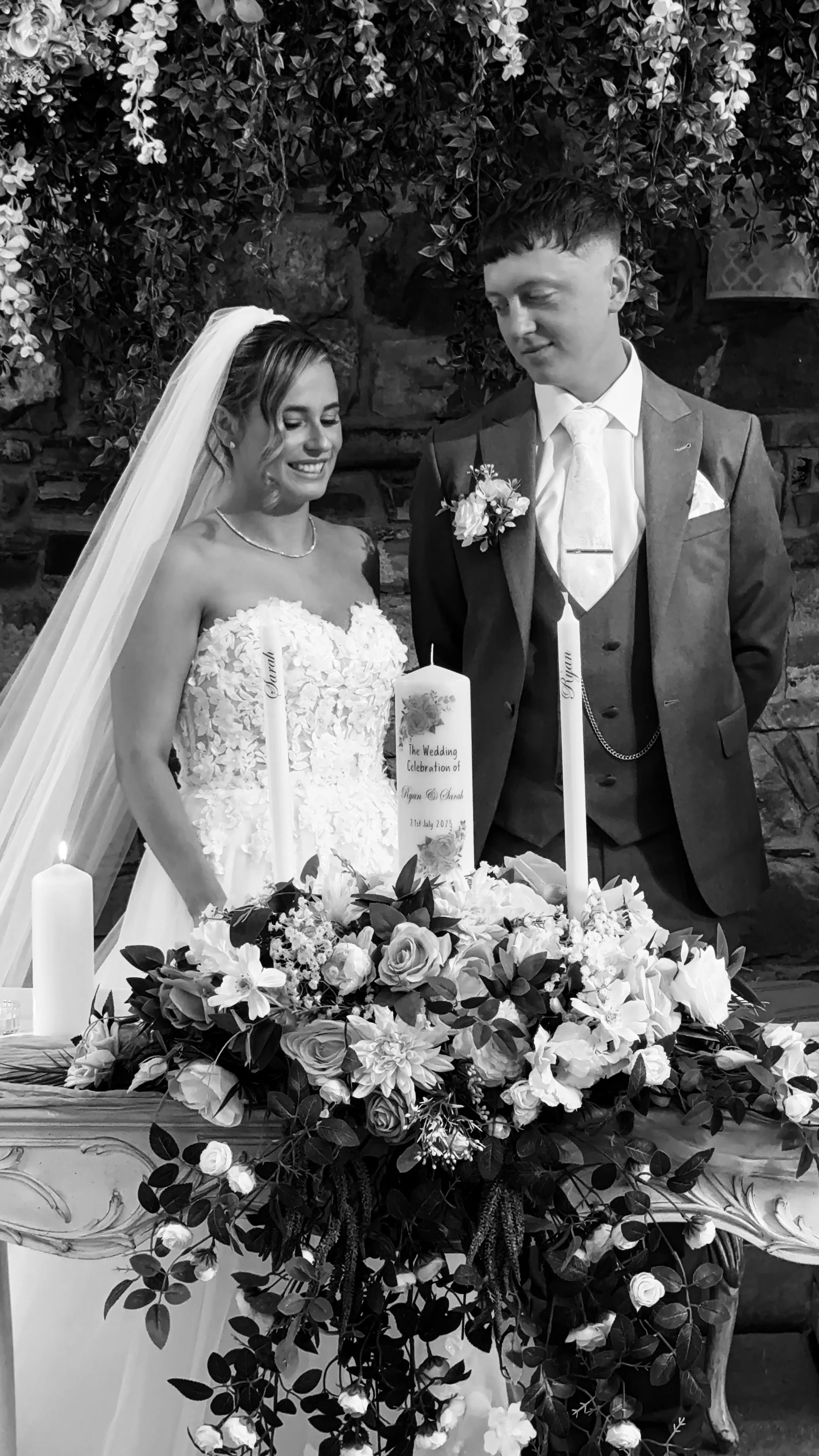 Black and white photo of a bride and groom standing behind a decorated table with flowers and candles during a wedding celebration.