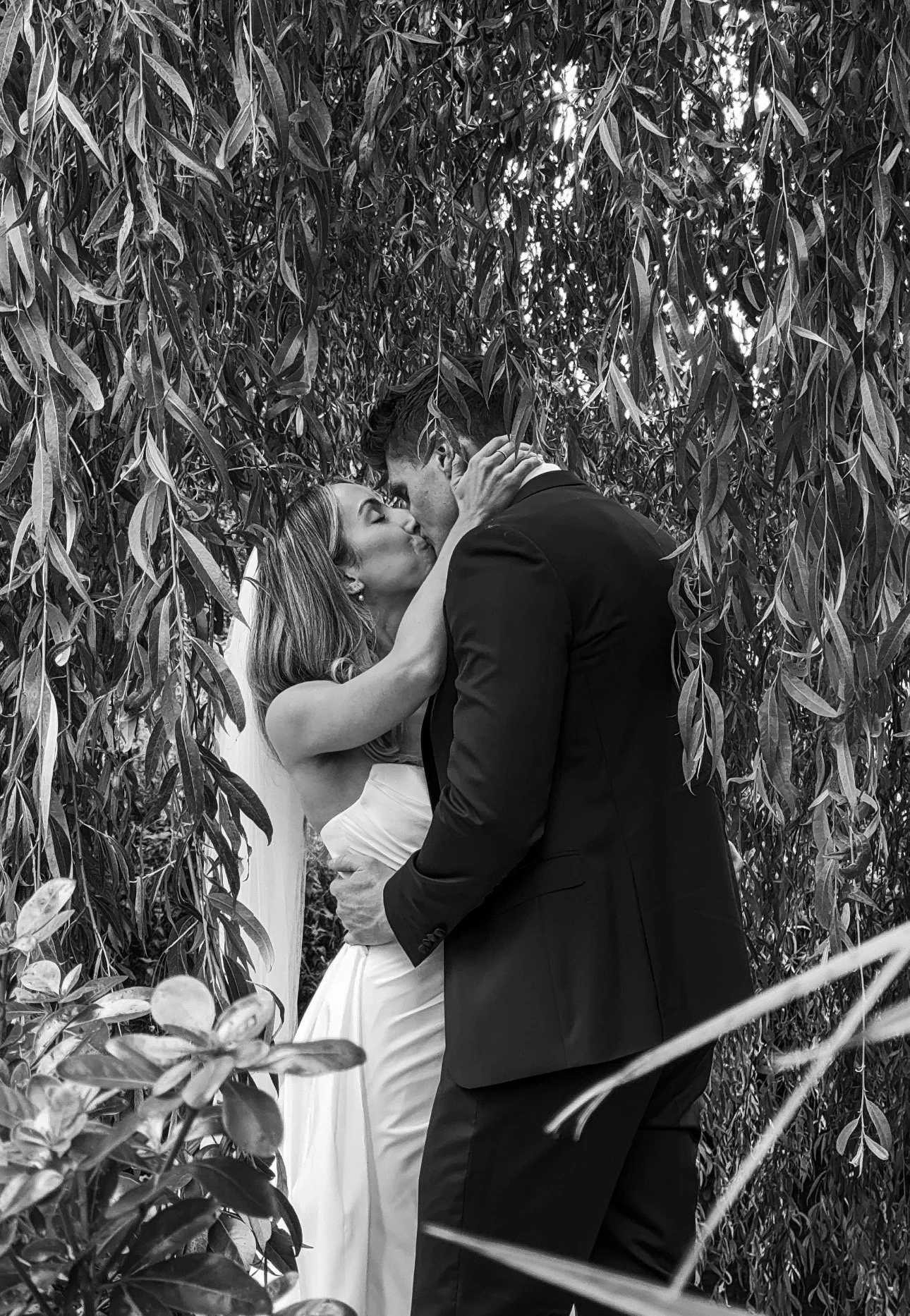 A black-and-white photo of a bride and groom kissing, surrounded by dense foliage, with the bride wearing a strapless white dress and the groom in a suit. Wedding Content Creator Ireland in Kilkea Castle, Kildare